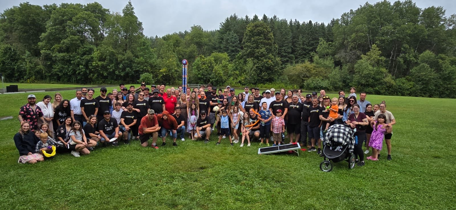 Group photo of diverse people, children, and families outdoors on a grassy field with trees in the background and a tall signpost, during daytime.