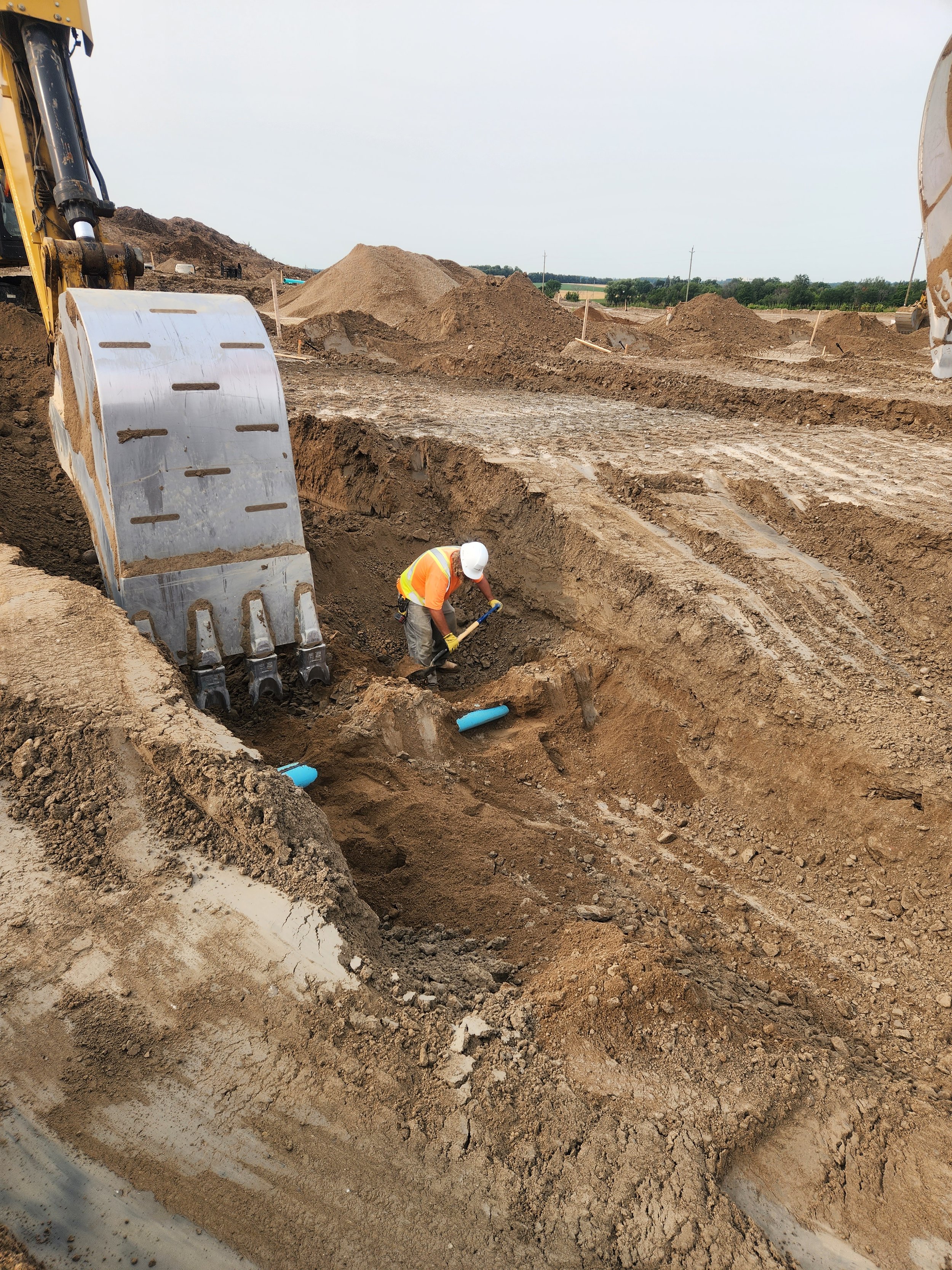 Construction worker in safety gear working on laying blue pipes underground at a construction site with earthmoving equipment and large dirt mounds in the background.