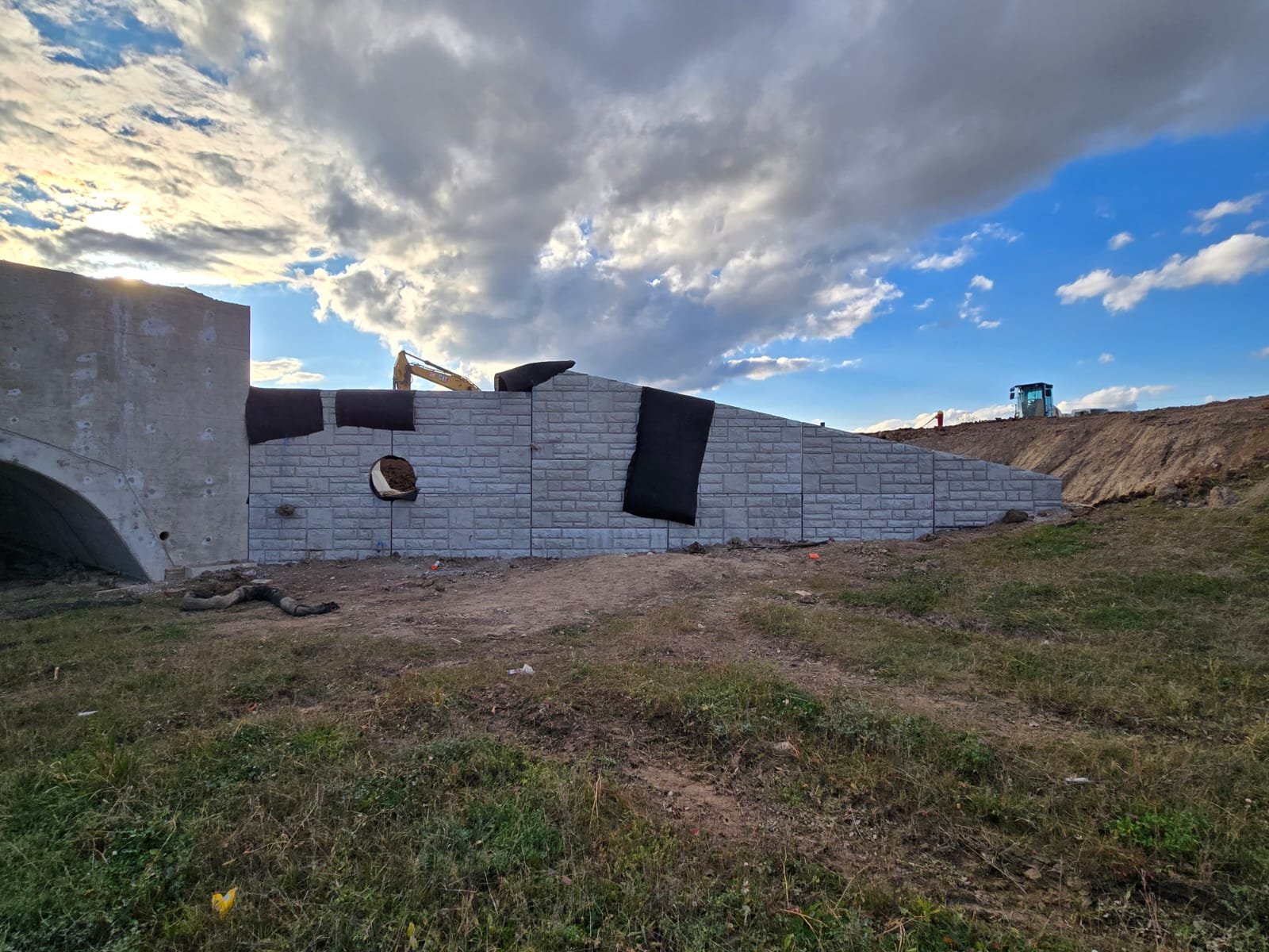 Construction site with a partially built retaining wall made of gray bricks, black waterproofing material on top, a construction excavator visible behind the wall, and a dirt road with patches of grass under a partly cloudy sky.