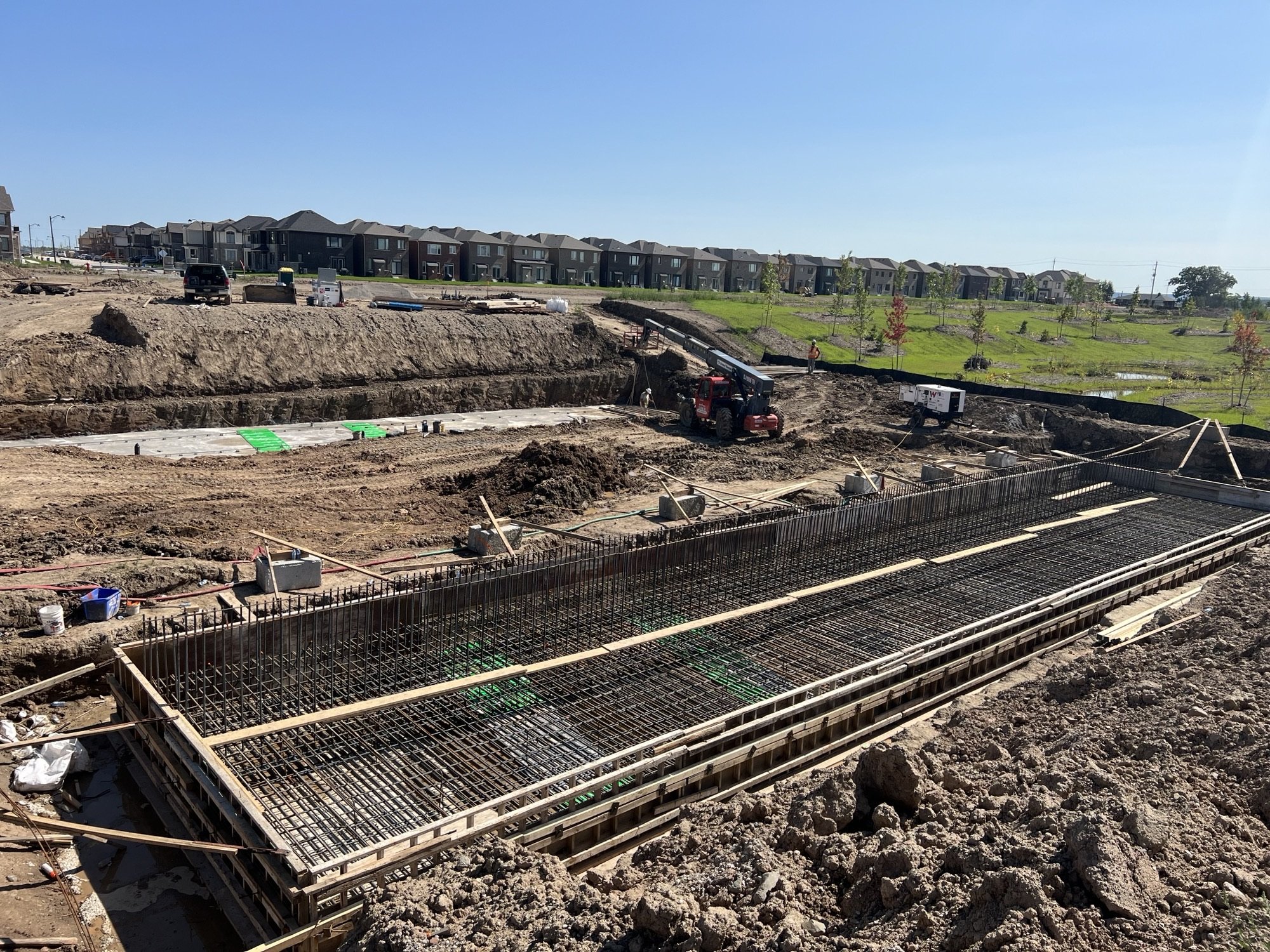 Construction site with steel rebar foundation in progress, a small crane, and construction workers, with residential houses and green trees in the background on a sunny day.