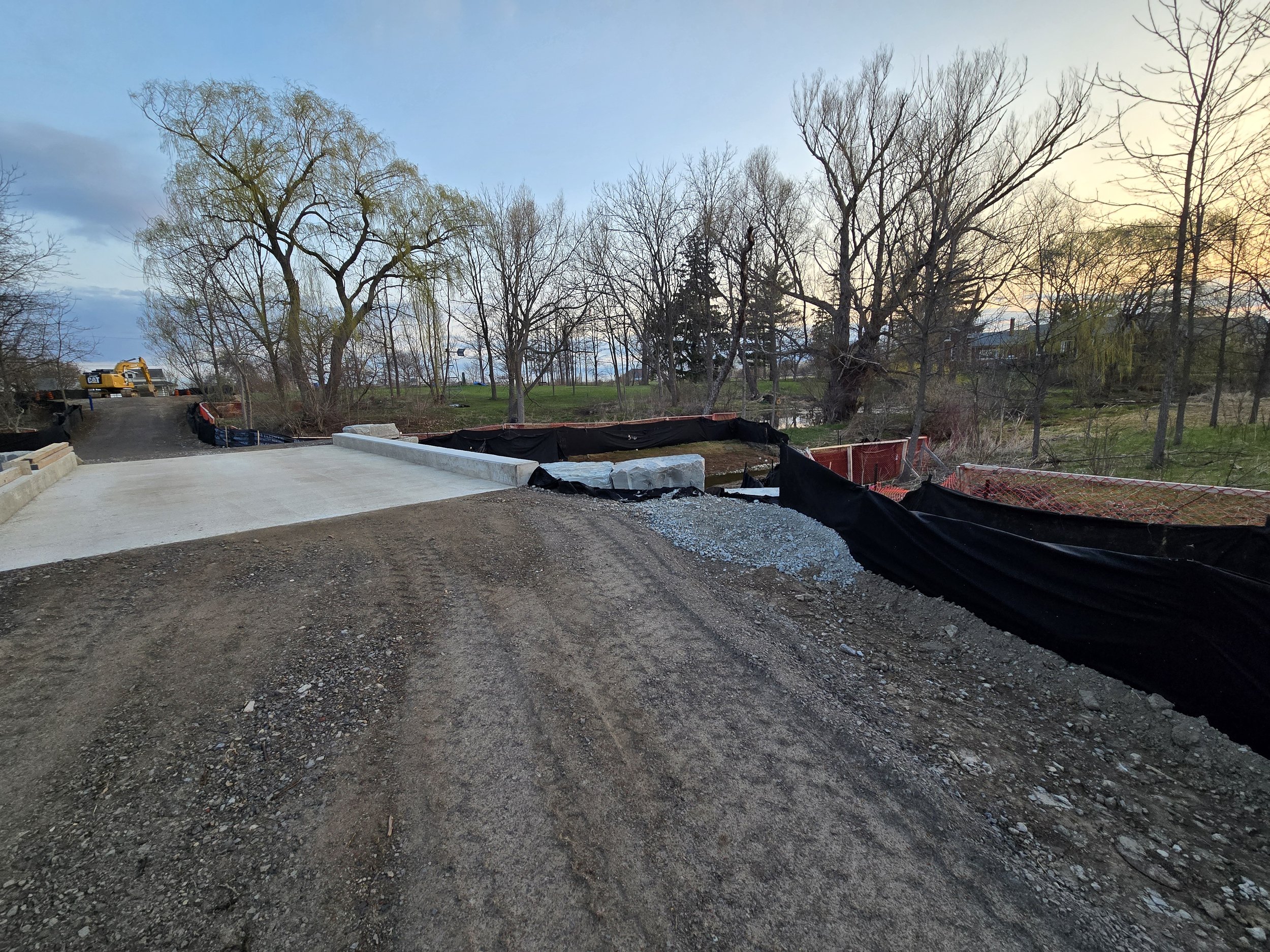 A construction site with a new concrete sidewalk and gravel, black setback fencing, a red safety barrier, trees with bare branches, and a sunset sky in the background.