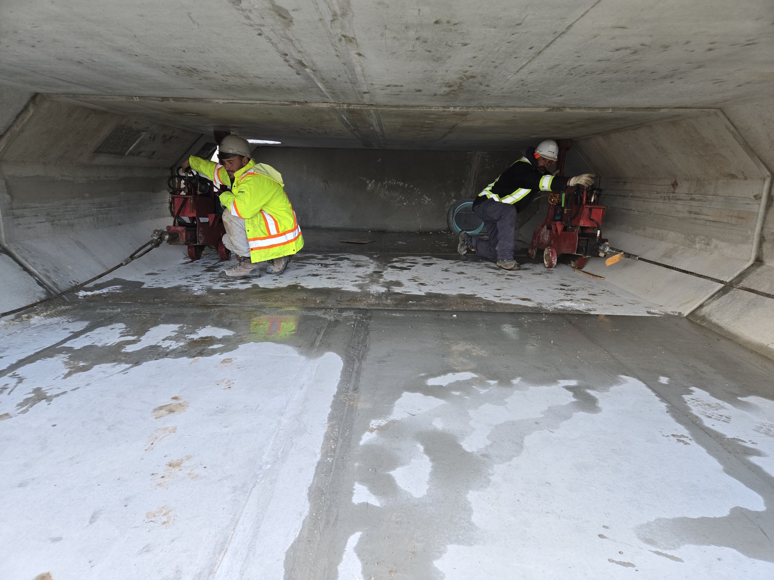 Two construction workers are operating power saws inside a concrete tunnel or underground passageway, working on the floor surface.