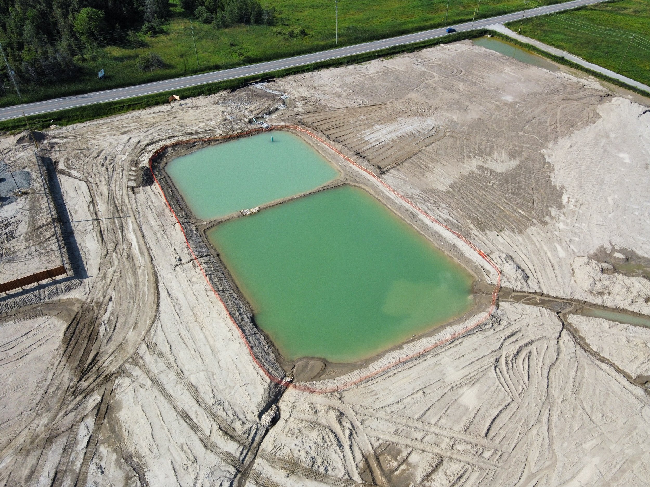 Aerial view of a construction site with two rectangular green ponds surrounded by dirt roads and land, near a highway with a few vehicles.
