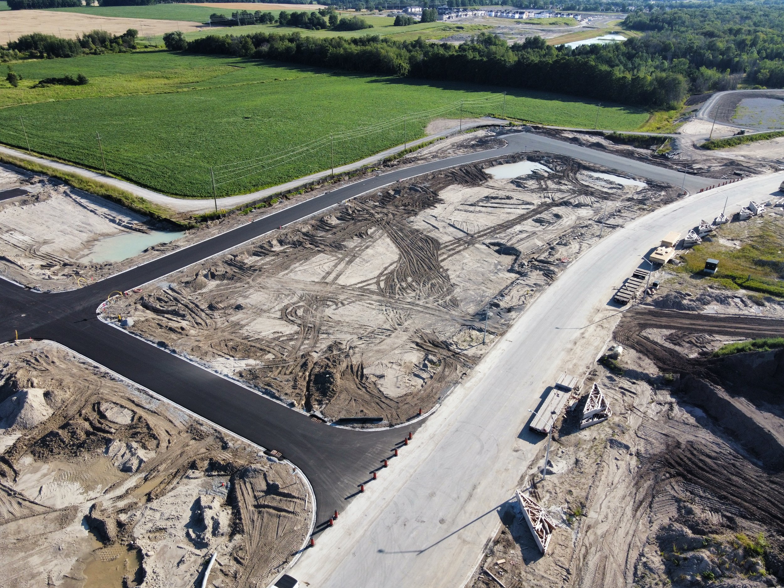 Aerial view of a construction site with newly paved roads, dirt pathways, and construction equipment, surrounded by green fields and trees.