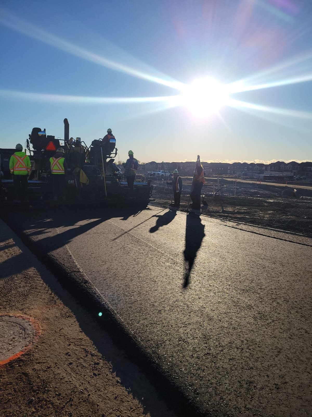 Construction workers wearing safety vests and helmets working on road construction under bright sunlight, with shadows cast on the newly paved road and houses in the background.