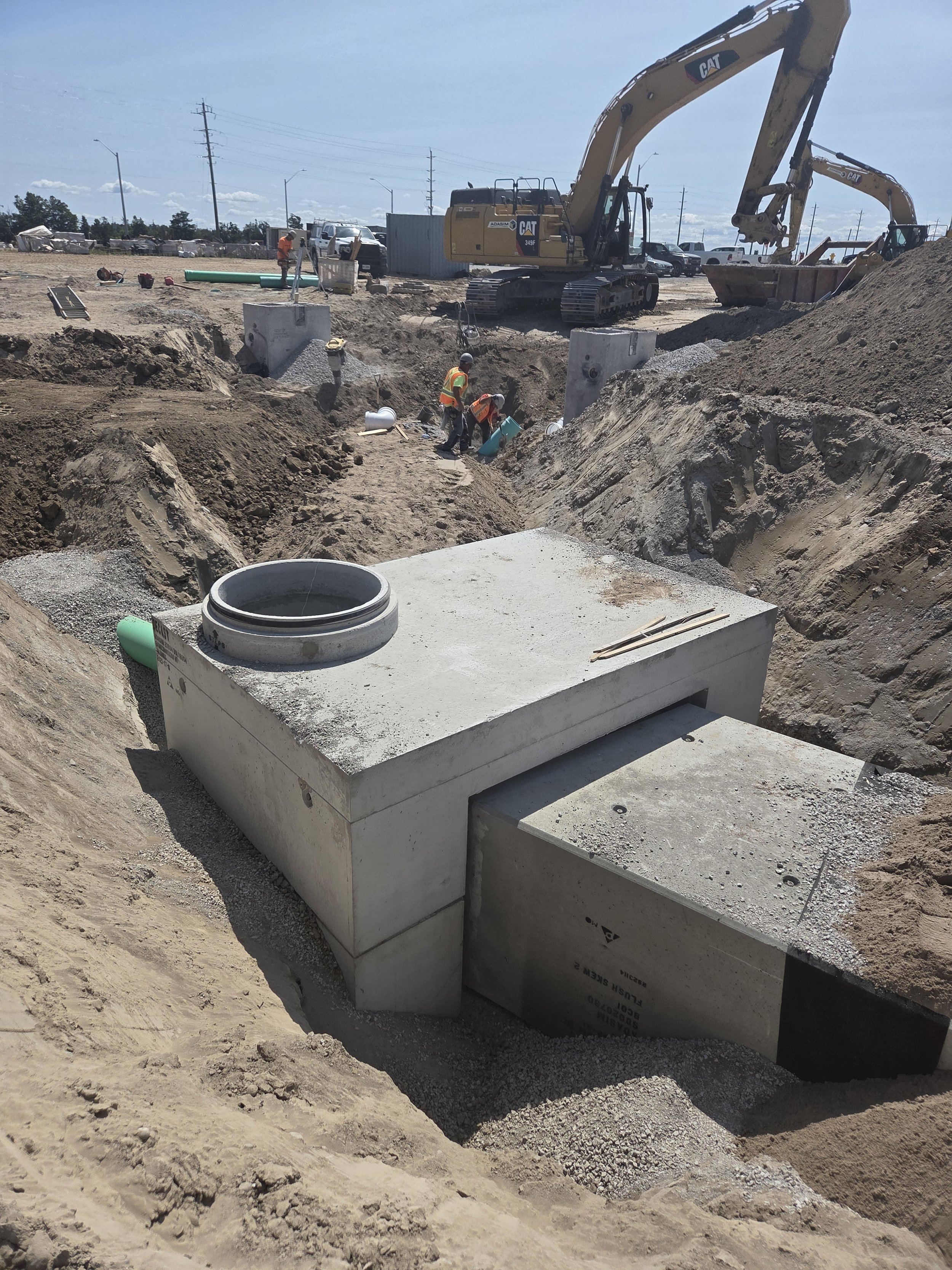 Construction site with workers and heavy machinery, including excavators, installing underground utility infrastructure with concrete structures and piping.