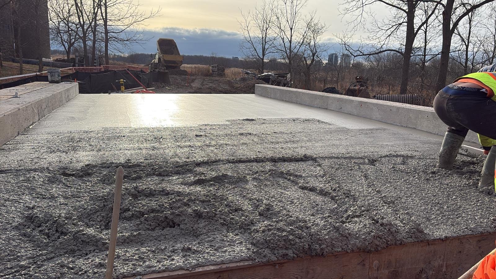 A construction worker in safety gear, installing a concrete sidewalk or pathway outdoors, with trees, construction equipment, and a cloudy sky in the background.