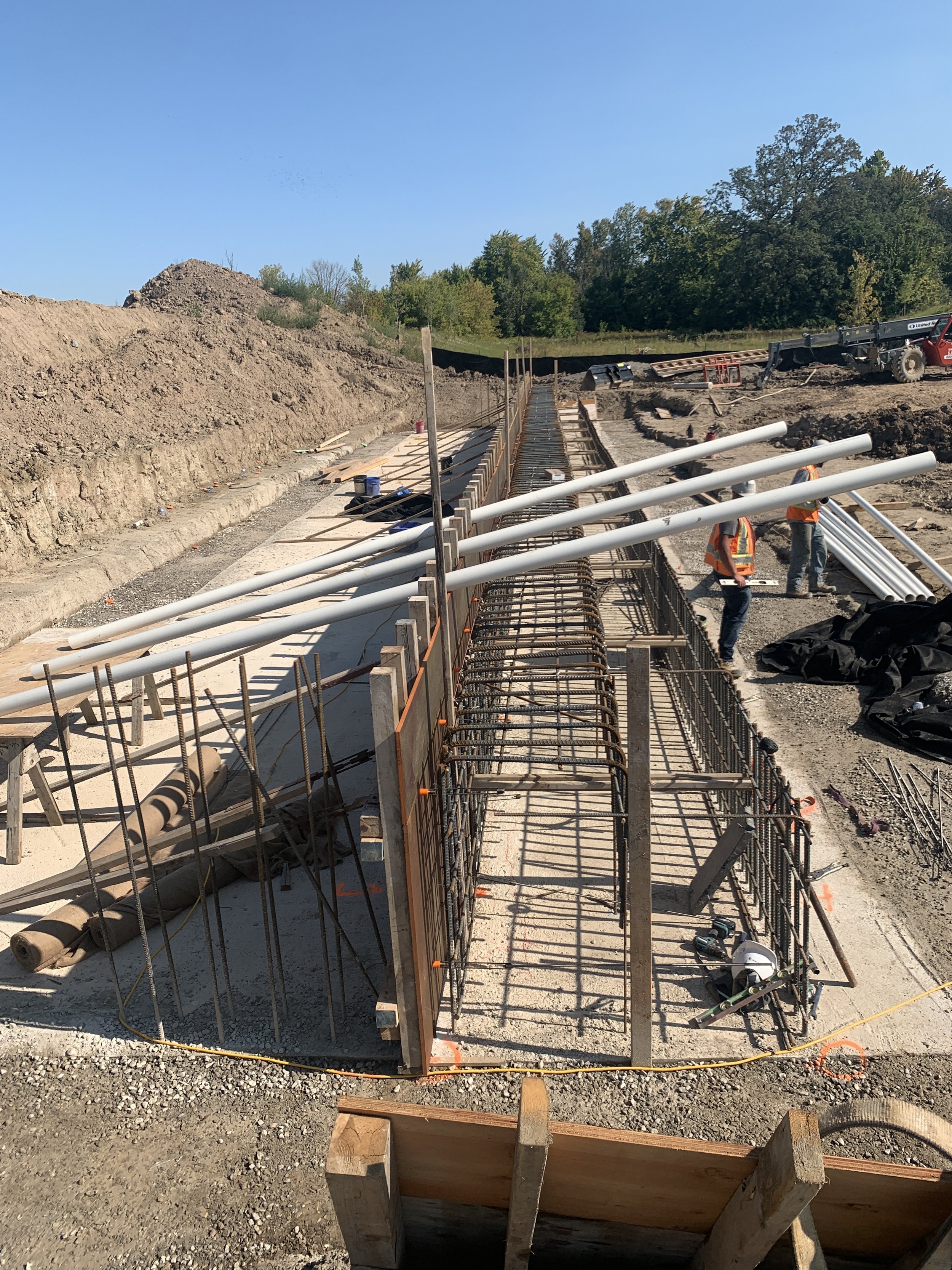 Construction workers building a concrete foundation with rebar and metal framing for a structure, with large PVC pipes laid across the site and a clear blue sky overhead.