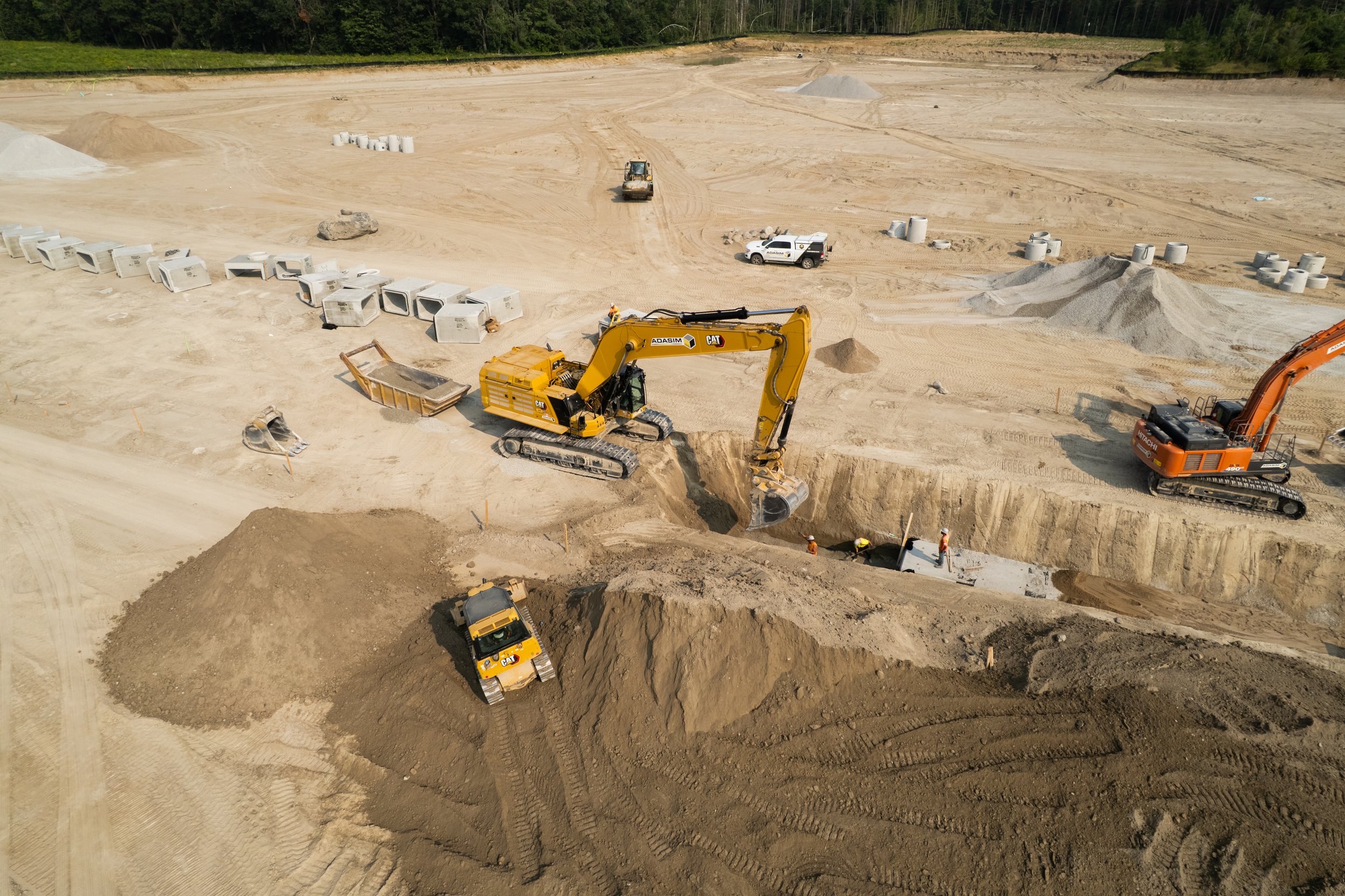 Construction site with heavy machinery, including excavators and a bulldozer, working on digging and moving dirt. Several workers are present, and various construction materials and equipment are scattered across the site.