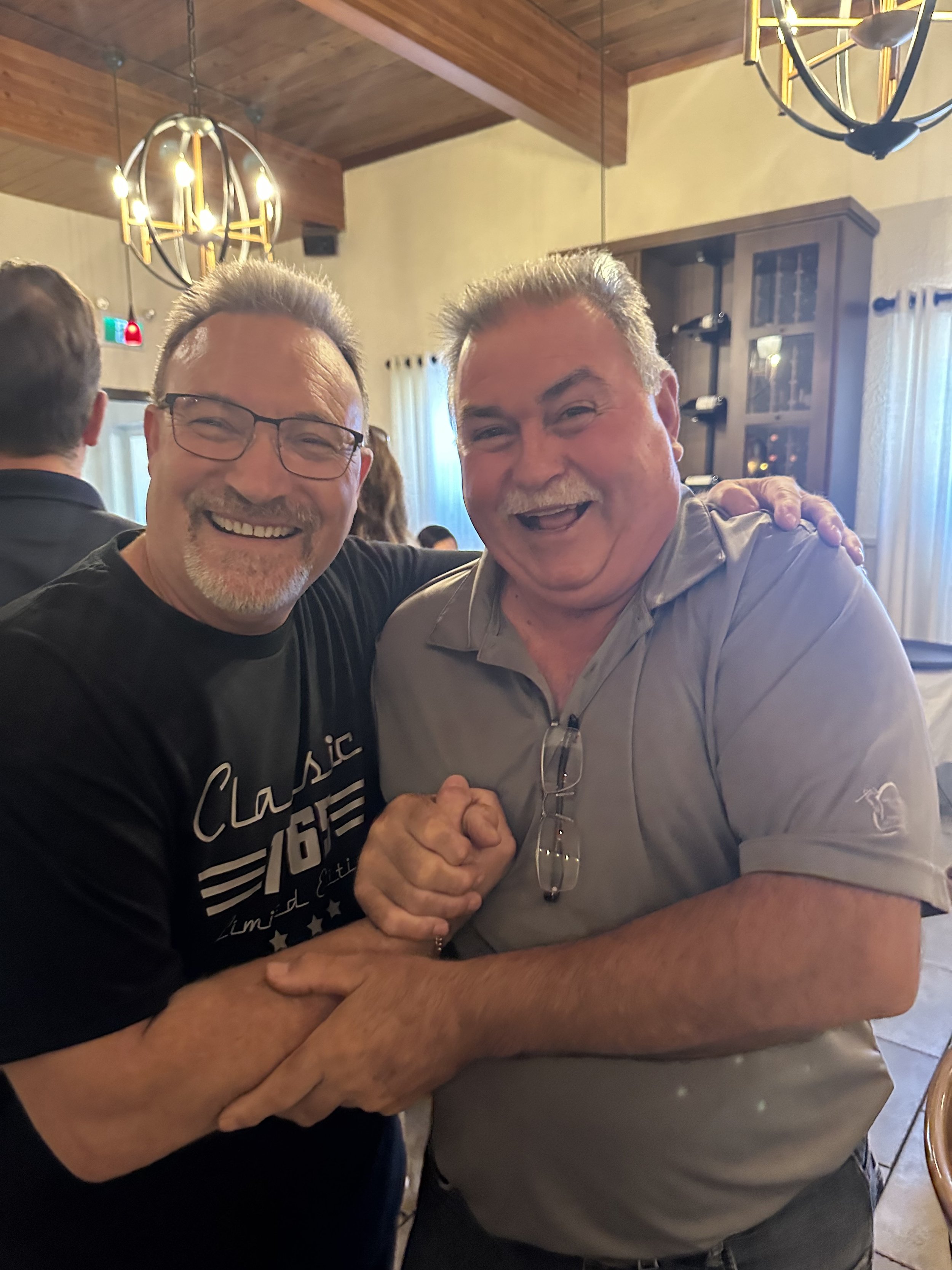 Two men smiling, shaking hands, and hugging in a warmly lit dining or event space with chandeliers, beige walls, and large windows with curtains.