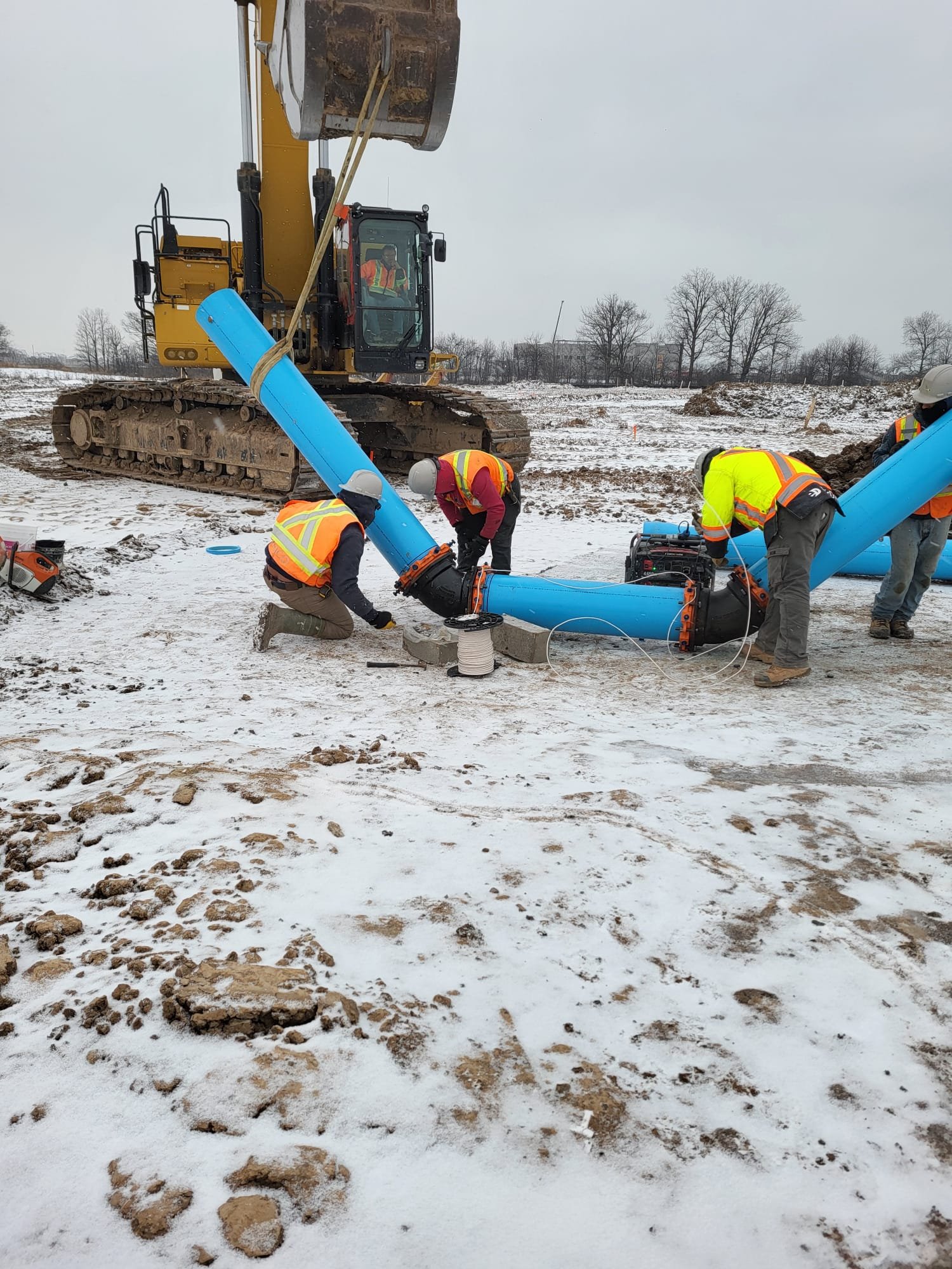 Workers in safety vests and helmets installing or repairing large blue pipeline pipes on a snowy construction site, with a tracked excavator in the background.