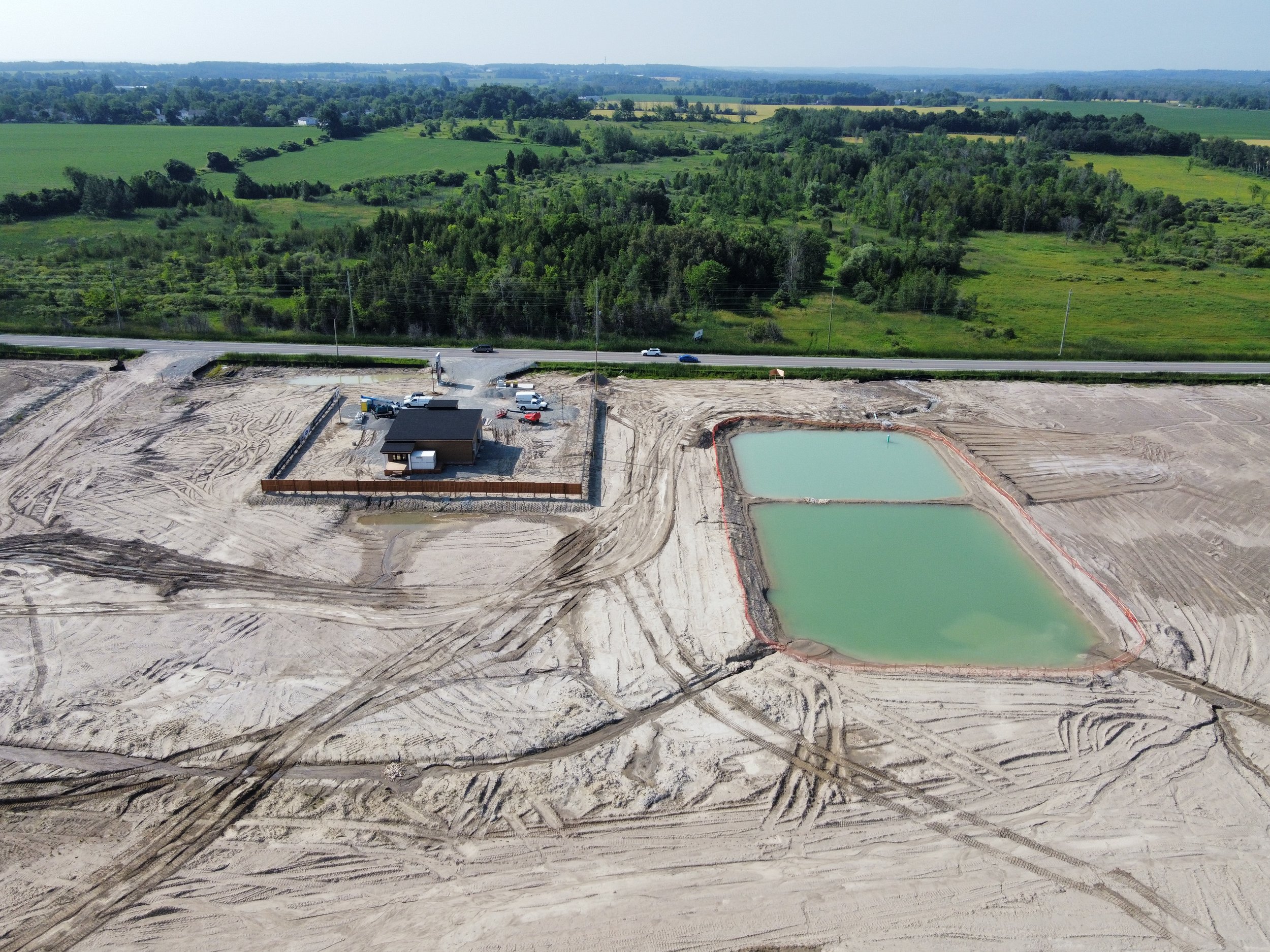 Aerial view of a construction site with a small building, surrounded by dirt roads, two rectangular green ponds, and a paved highway with vehicles passing by. Green fields and forested areas are visible in the background.