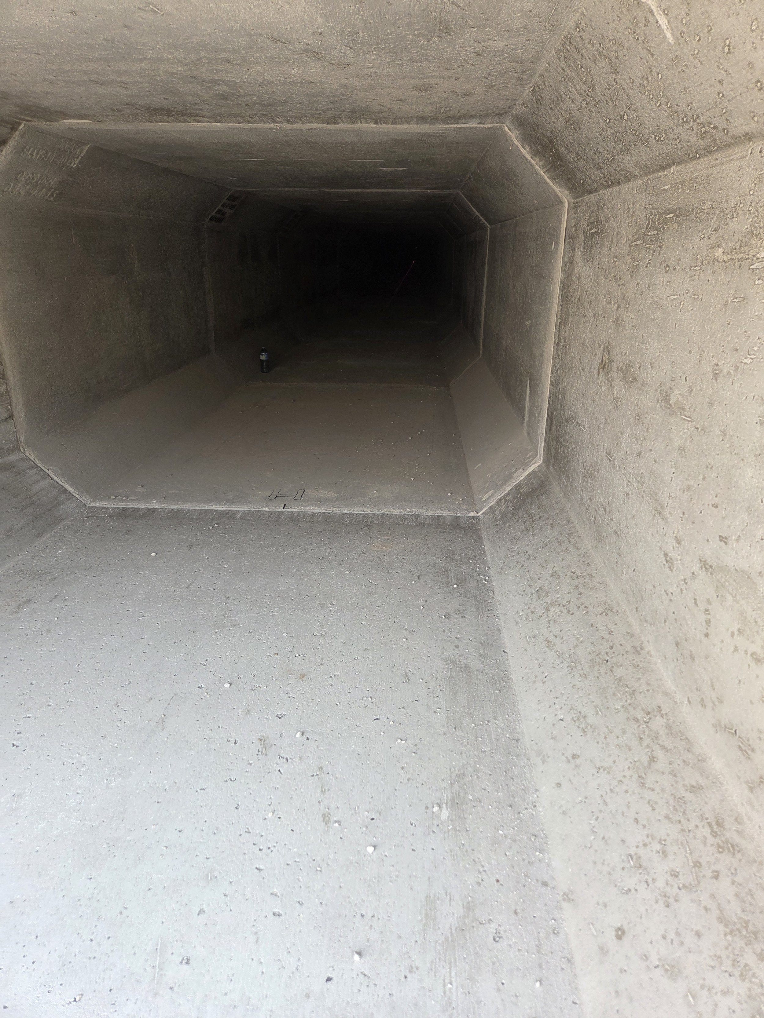 Looking into a dark, concrete industrial duct or tunnel with a water bottle placed on the floor near the entrance.