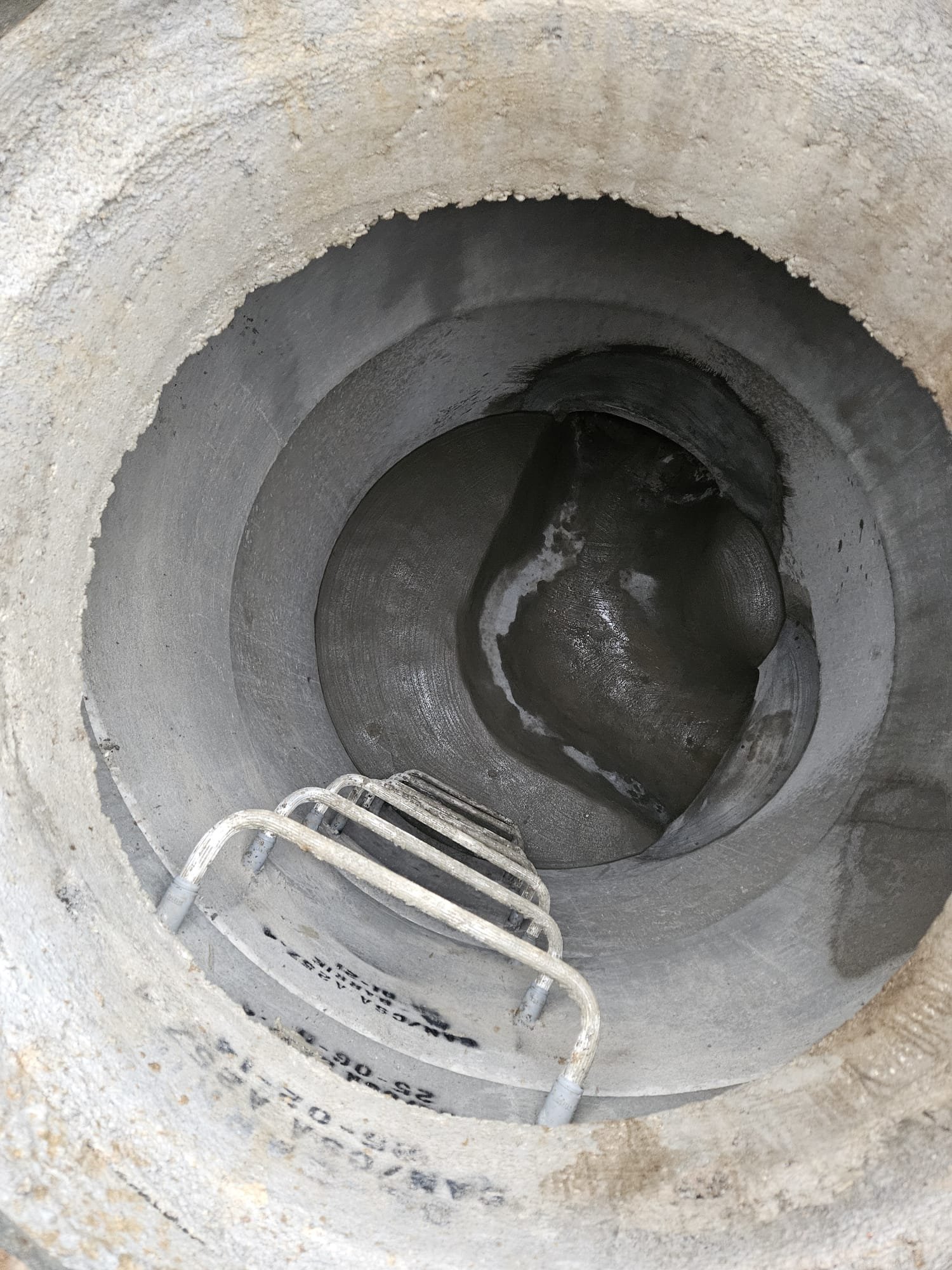 The photo shows the inside of a cylindrical concrete structure, with a metal ladder attached on one side, leading downward into the dark space.