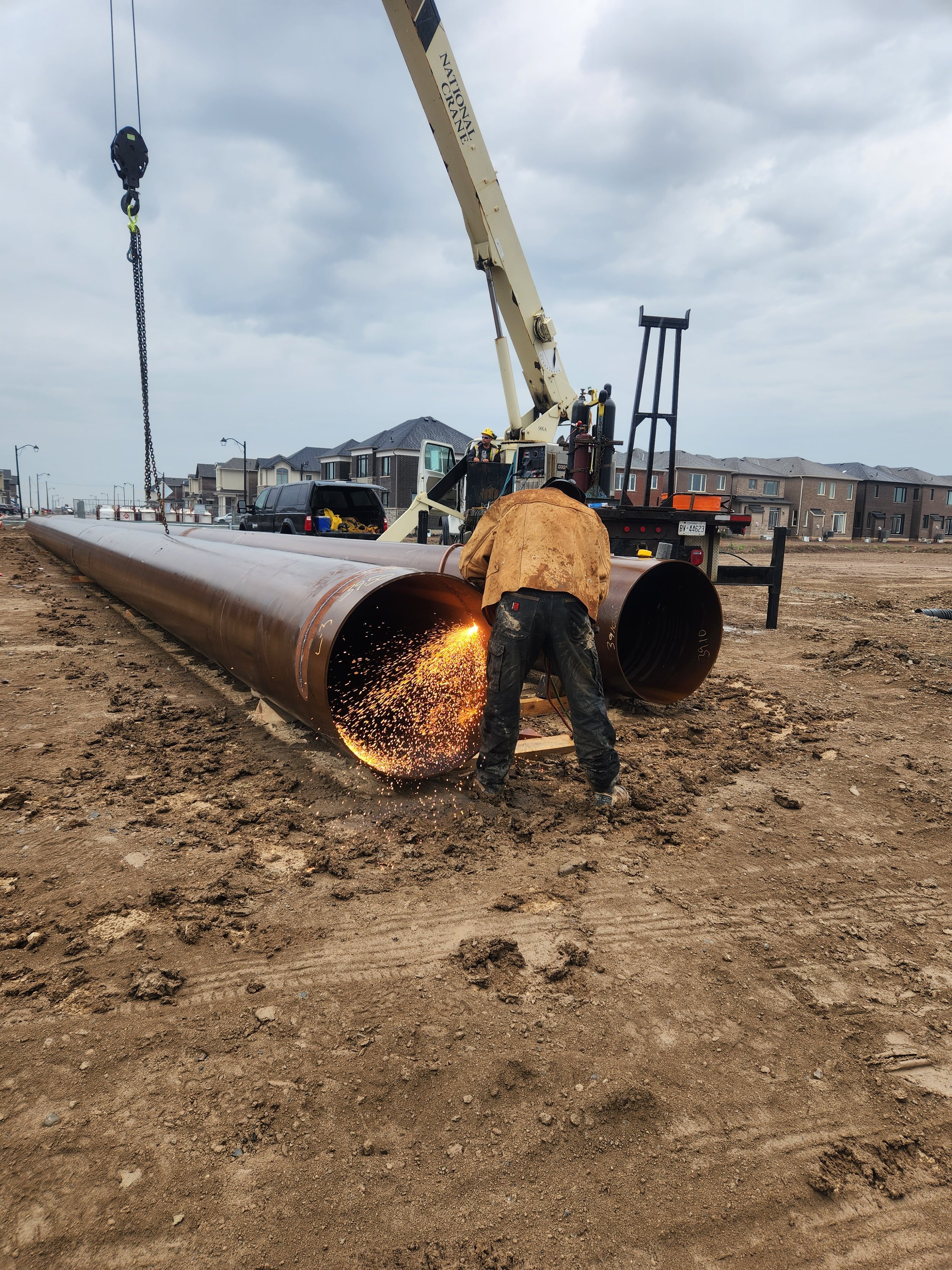 A construction worker welding a large pipe at a construction site with a crane overhead and a residential neighborhood in the background.