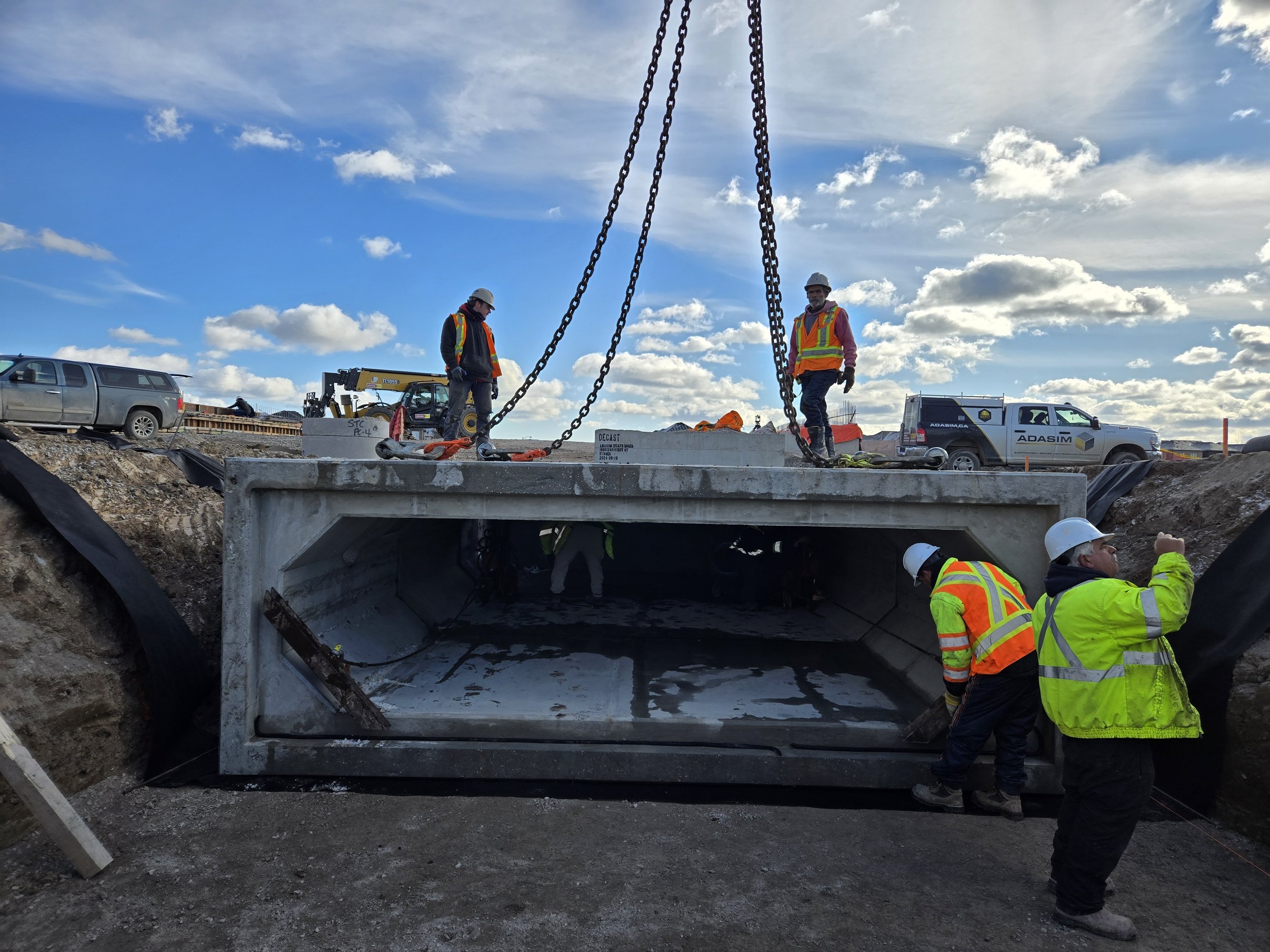 Construction workers installing a concrete culvert under a roadway, with equipment and vehicles in the background, under a partly cloudy sky.