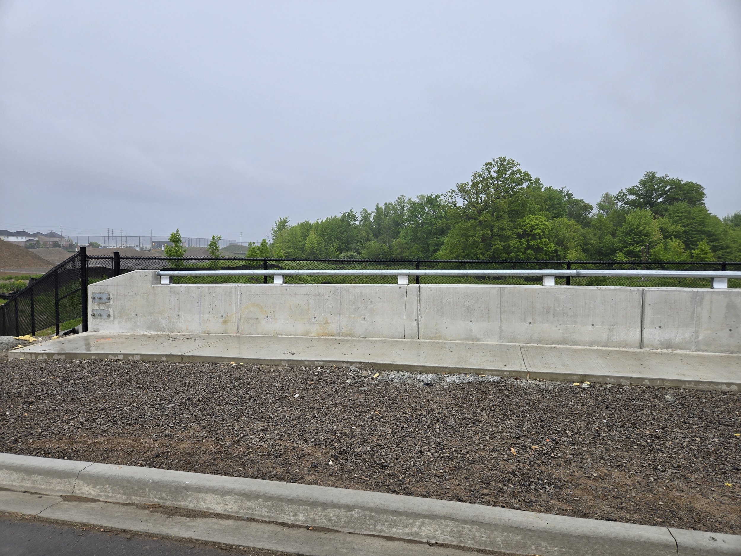 Concrete barrier on a roadside with safety fencing and green trees in the background under an overcast sky.