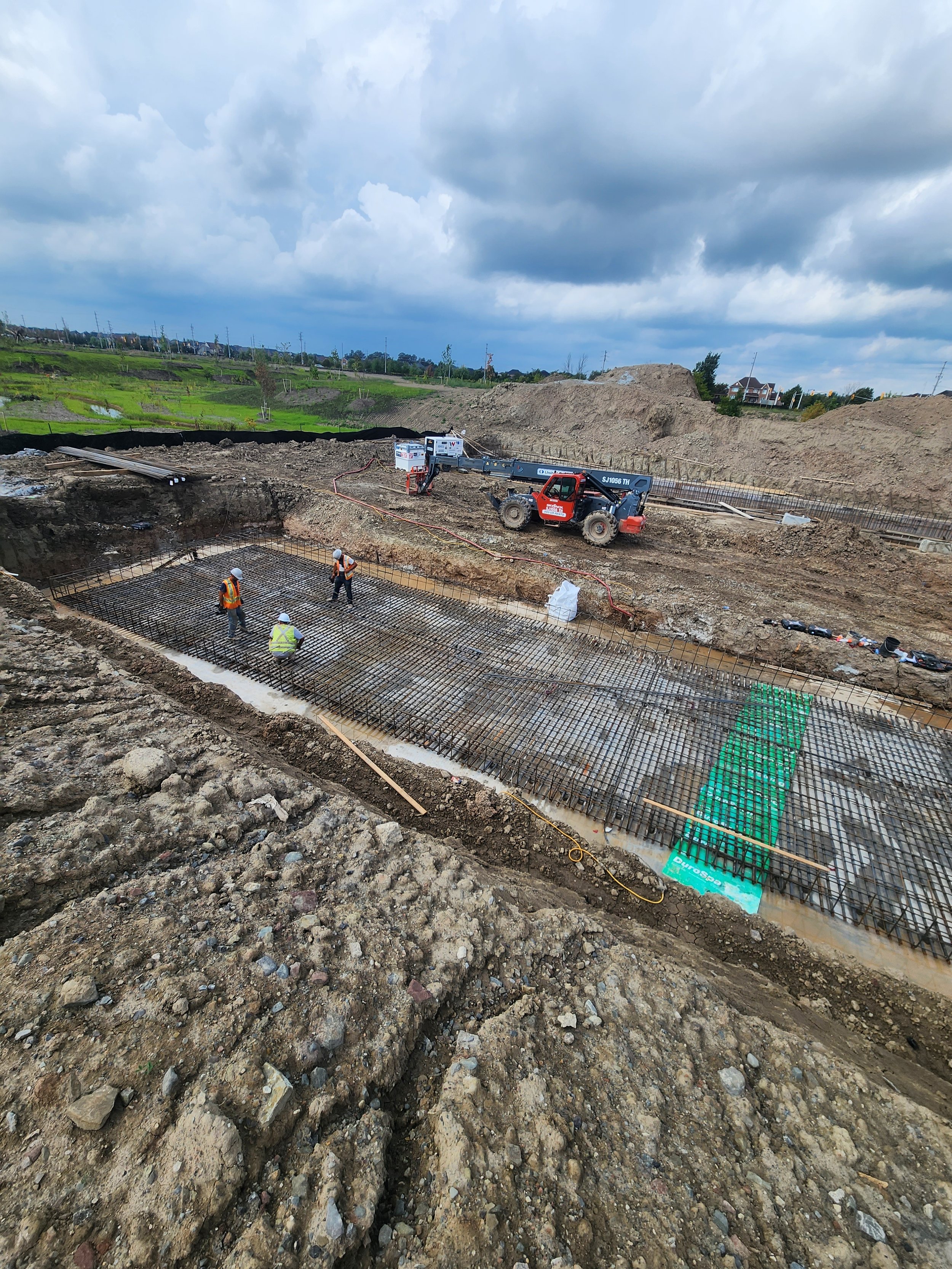 Construction site with workers laying rebar for a foundation, heavy machinery, and a cloudy sky in the background.
