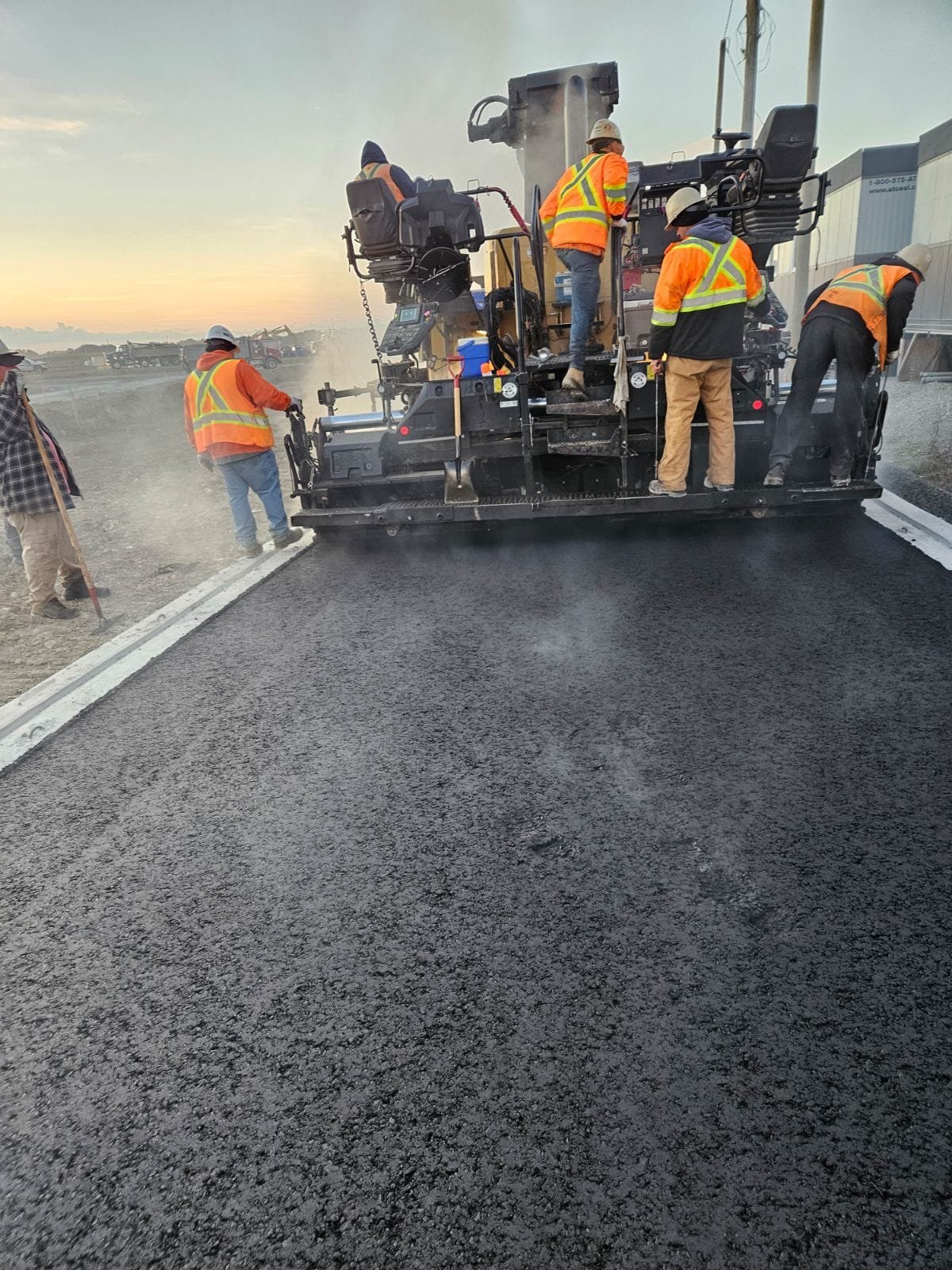 Construction workers wearing orange safety vests and white helmets operating a large asphalt paving machine on a highway at sunset.
