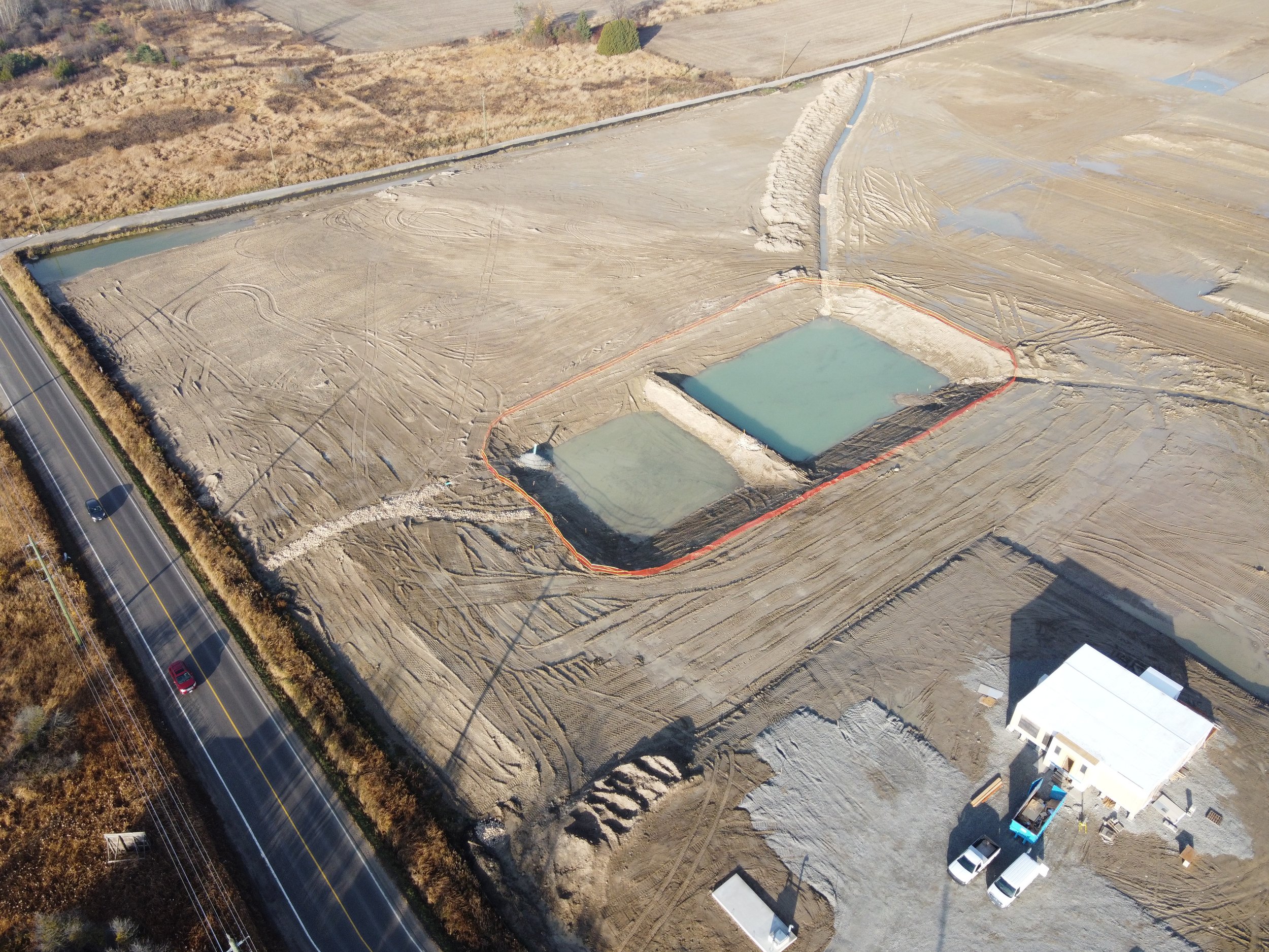 Aerial view of a construction site with two large rectangular water ponds and a small white building nearby. The site is surrounded by dry land and a road with cars.