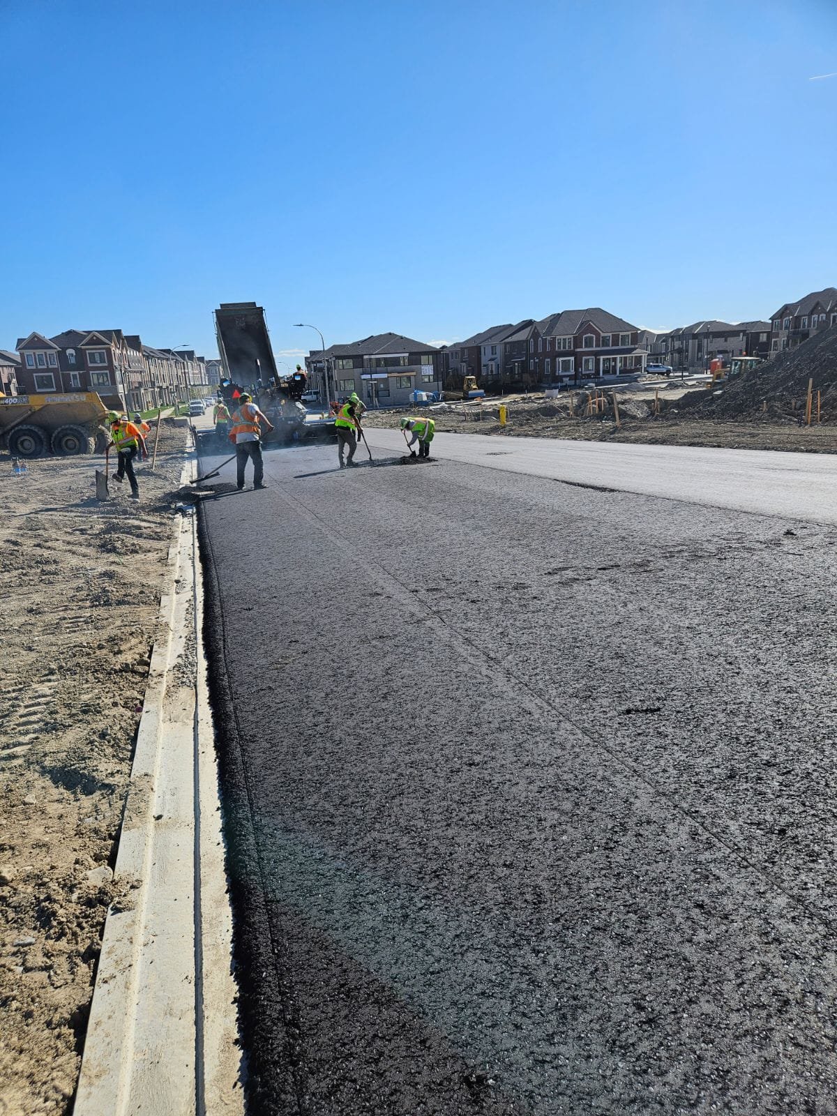 Workers in safety vests are paving a road in a residential area under a clear blue sky.