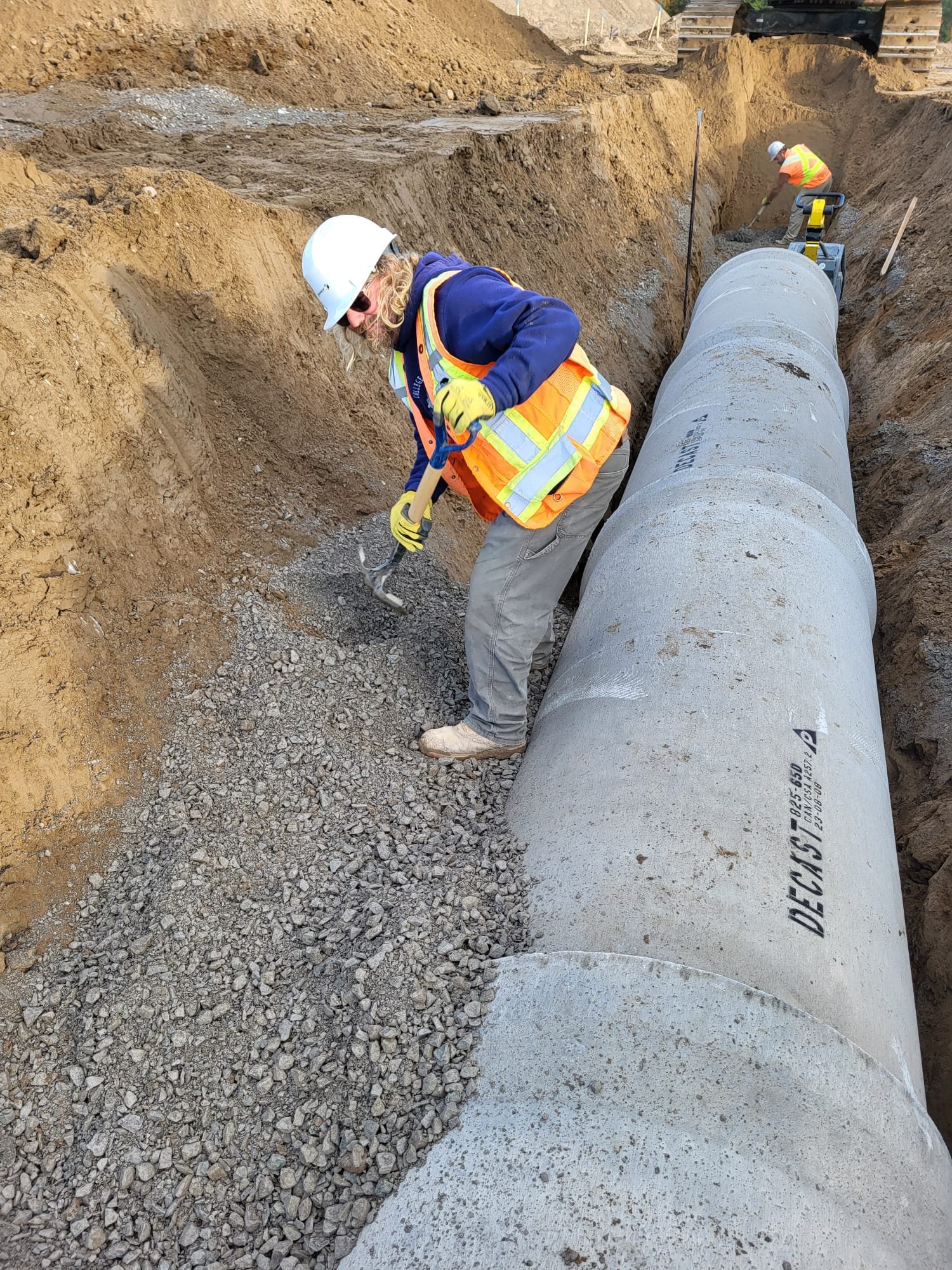 Construction workers in safety gear working on a large concrete pipe installation in a trench, with one worker using a pickaxe and another working further back.