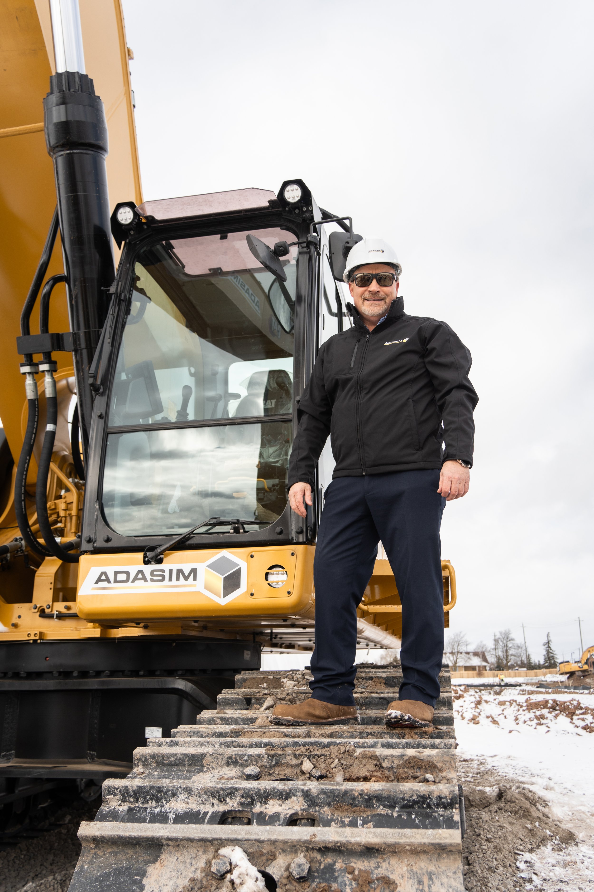 A man wearing sunglasses, a white safety helmet, and black outdoor clothing standing next to a yellow excavator at a construction site with snow on the ground.