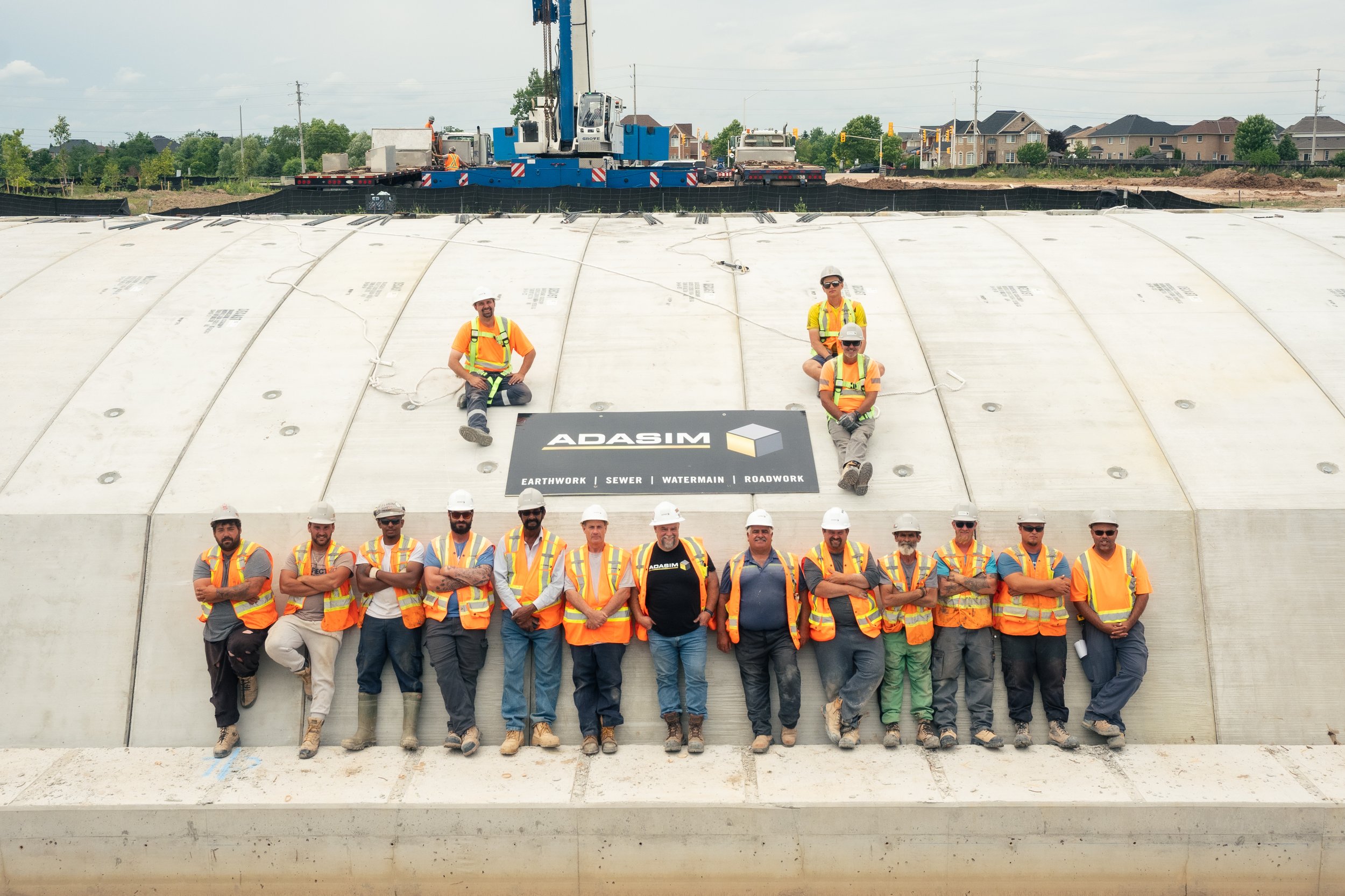 Group of construction workers in safety vests and helmets posing in front of a large concrete wall with the ADASIM logo, during daytime at a construction site with some vehicles and houses visible in the background.