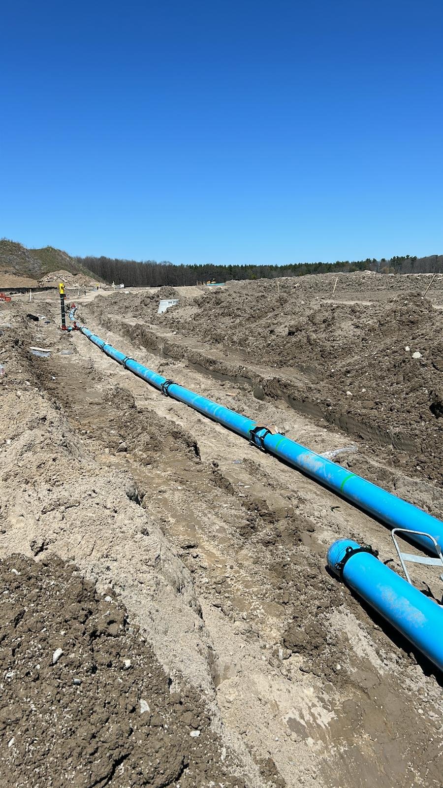 Construction site with blue piping laid across a trench in an open area under a clear blue sky.