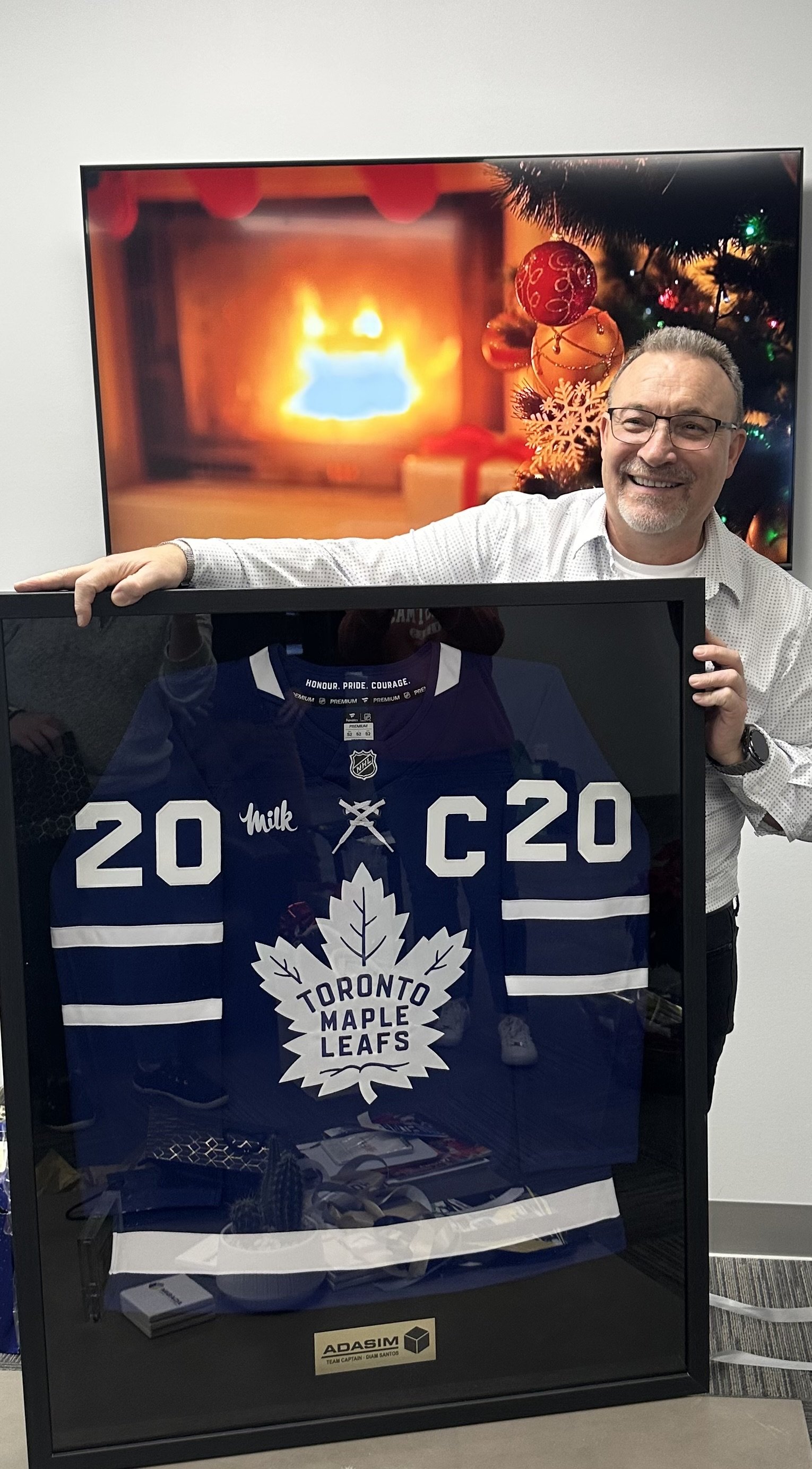 A man smiling and wearing glasses, holding a framed Toronto Maple Leafs hockey jersey with the number 20, in front of a white wall with a digital screen showing a cozy fireplace and Christmas decorations.