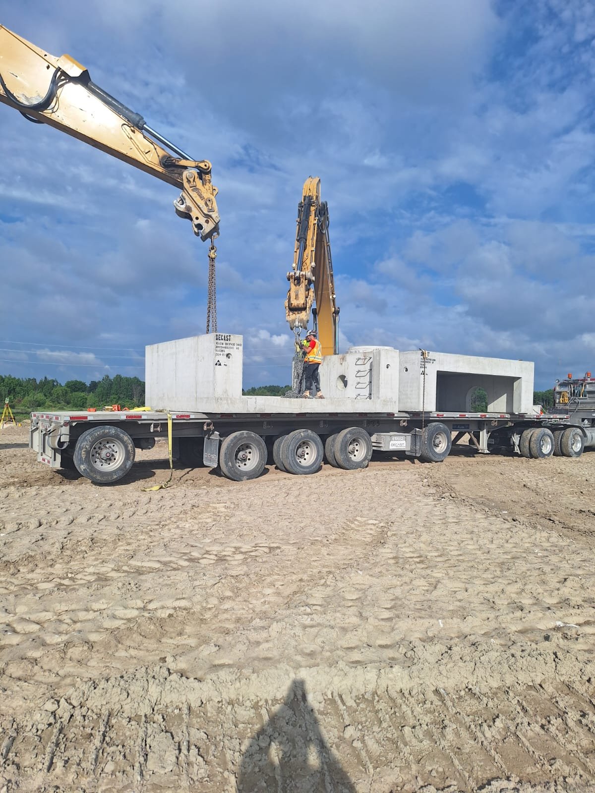 Construction workers operate a crane on a flatbed truck to place large concrete structures during a construction project at a dirt site under a partly cloudy sky.