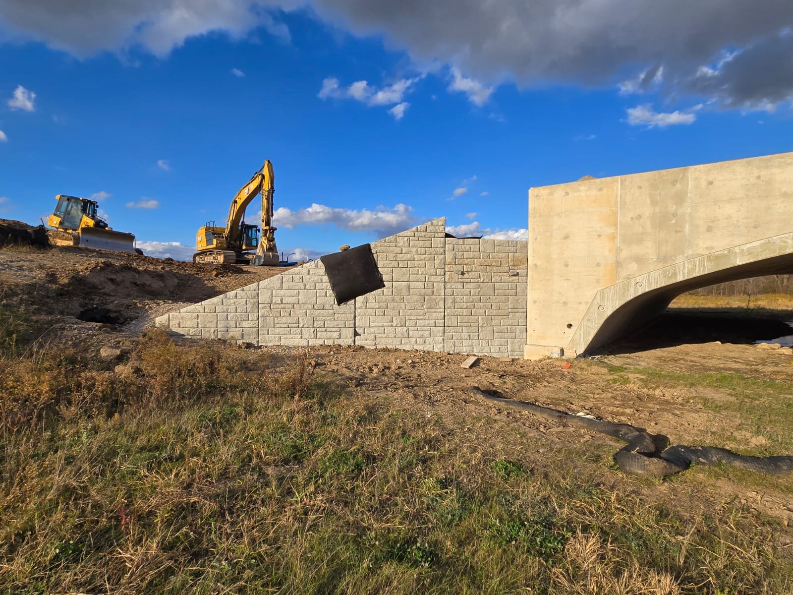 Construction site with two yellow excavators on a dirt hill, a concrete bridge with a brick barrier, and a partly cloudy blue sky.
