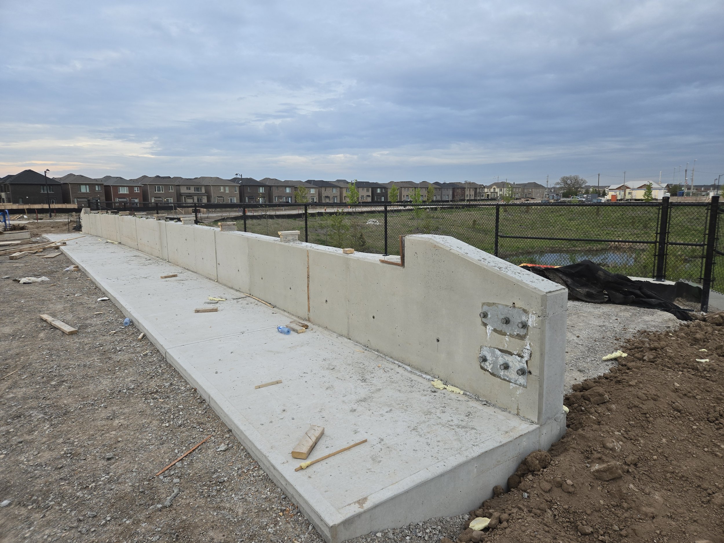 Construction site with a concrete barrier and sidewalk, overcast sky, residential houses in the background, black fence and a small pond.