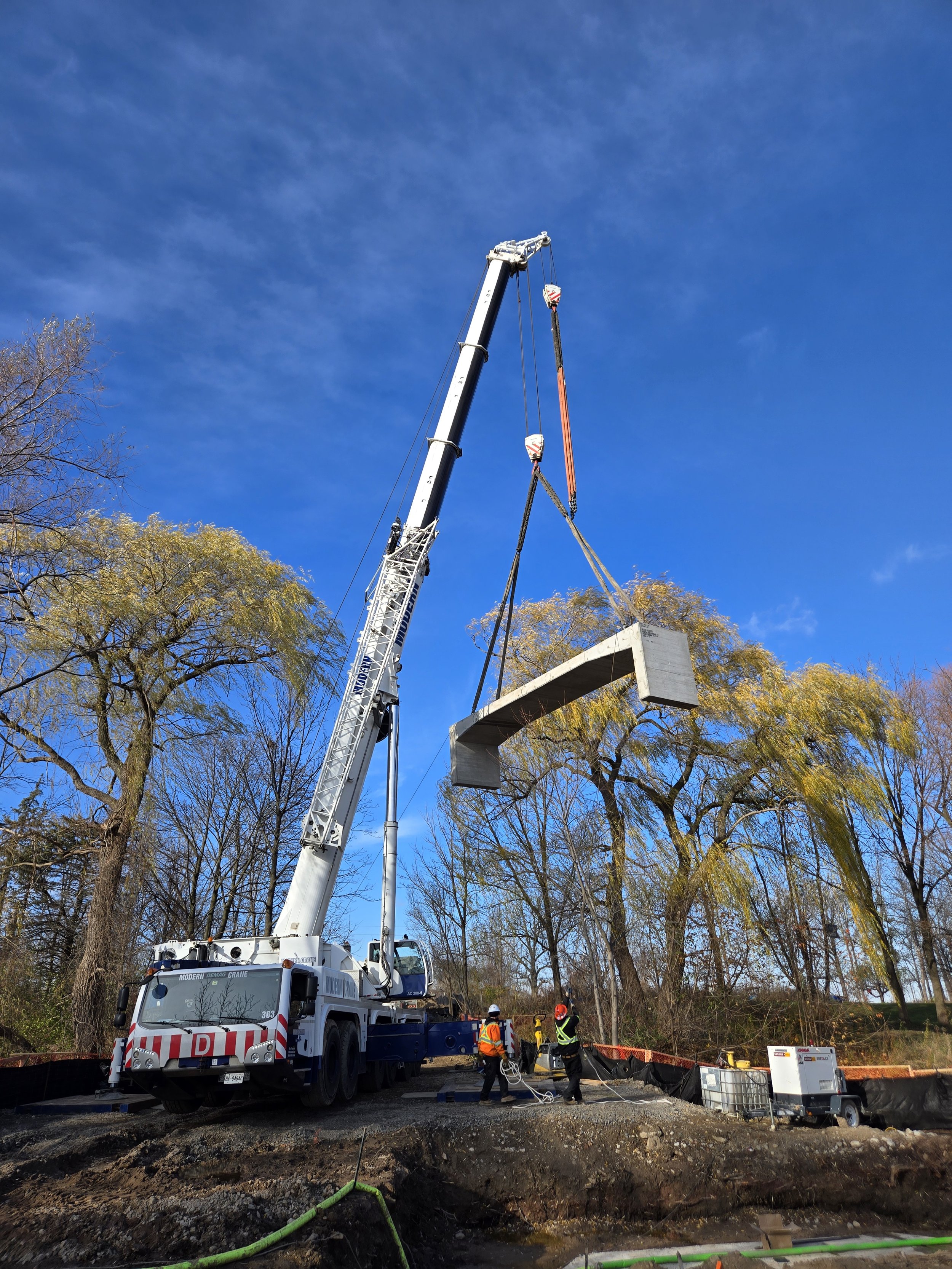 Construction workers guiding a large crane that is lifting a concrete bridge section at a construction site with trees and a blue sky in the background.