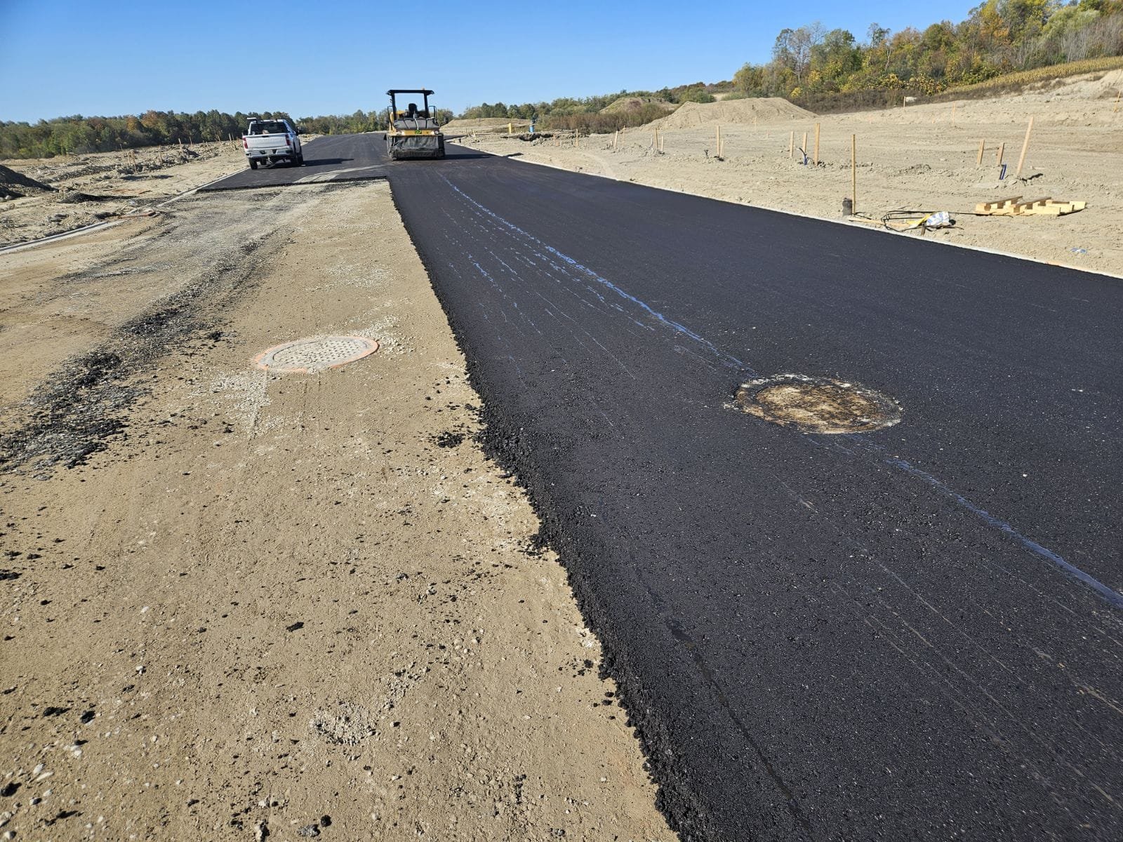 Construction workers are paving a new asphalt road with a bulldozer, a pickup truck, and road construction equipment visible in the background.