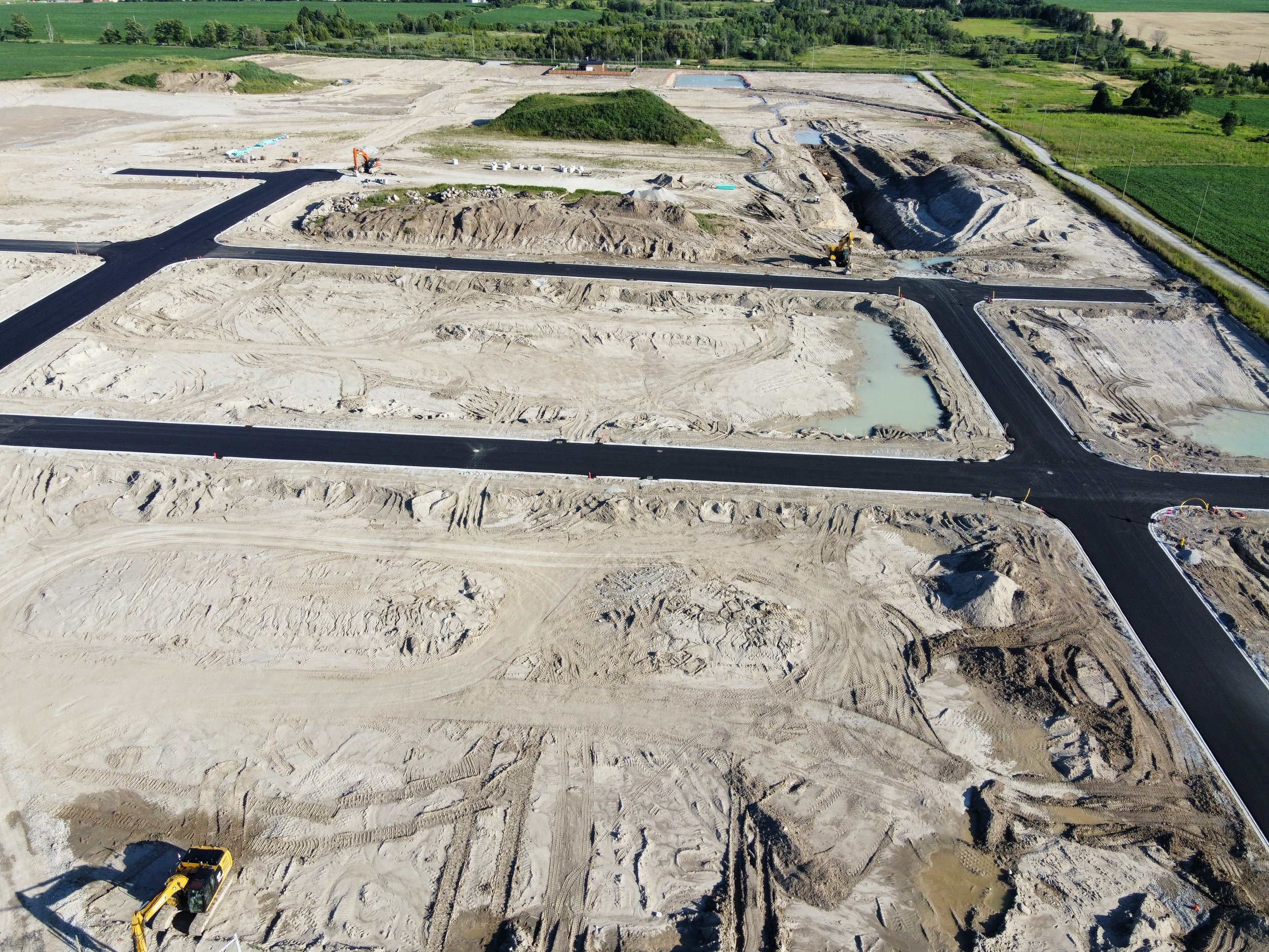 An aerial view of a construction site with new roads being paved amidst dirt and gravel, water puddles, and construction machinery, with green fields surrounding the site.