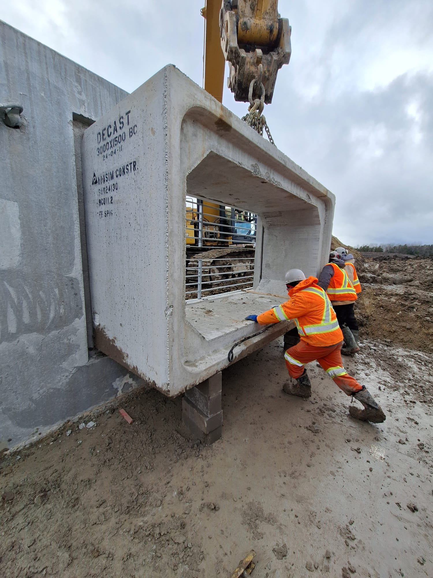 Construction workers in safety gear installing or positioning a large concrete structure at a construction site, with heavy machinery in the background.