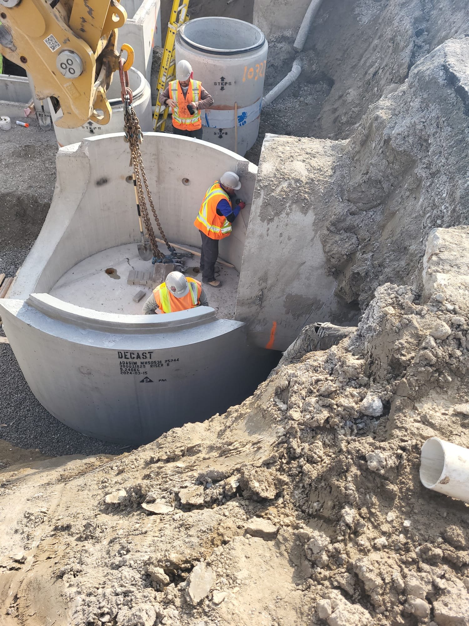 Construction workers in safety vests and helmets assembling a large concrete pipe into an underground infrastructure project, with construction equipment and additional pipes in the background.