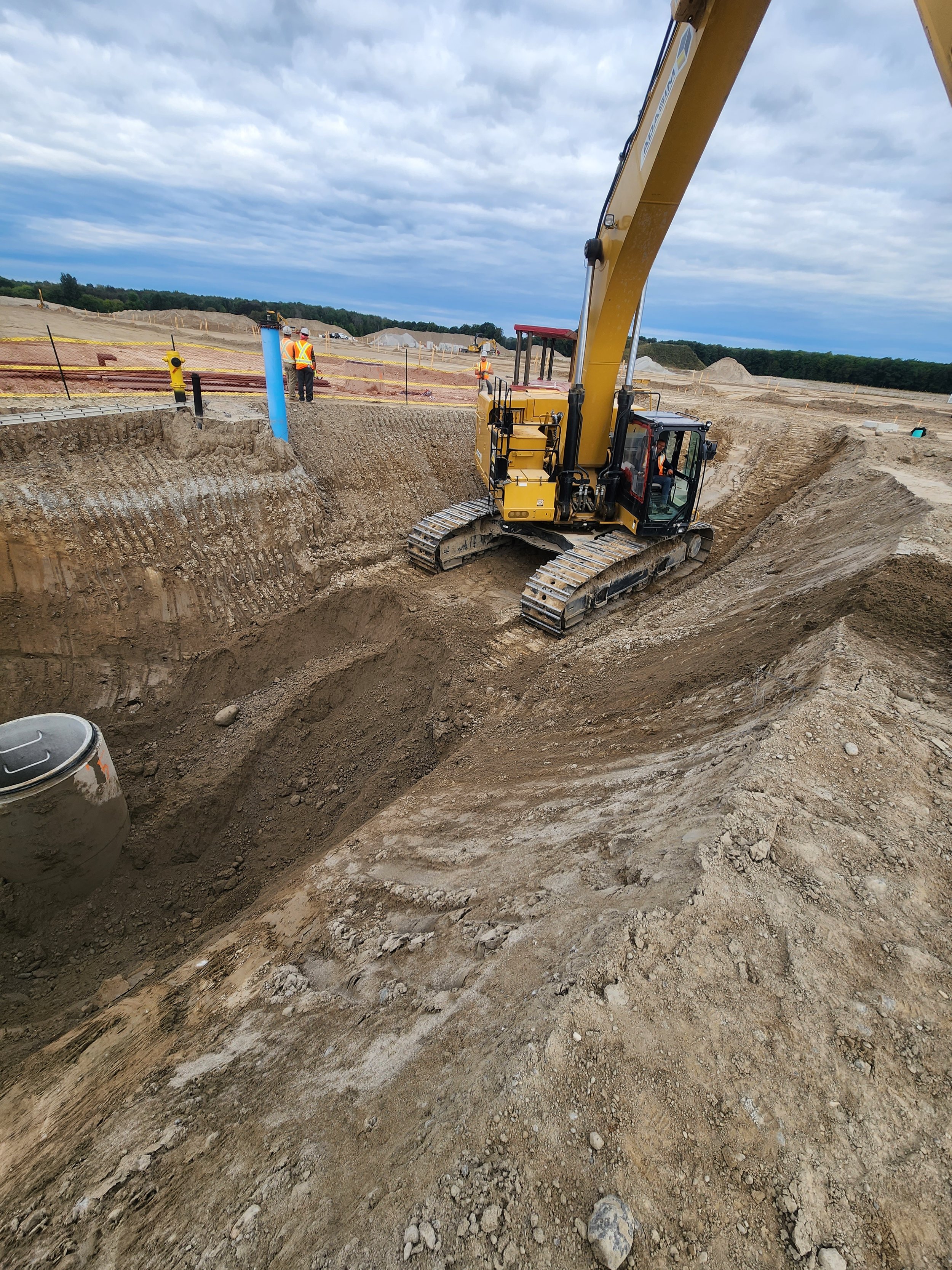 Excavator digging a trench at a construction site on a cloudy day with workers in the background.
