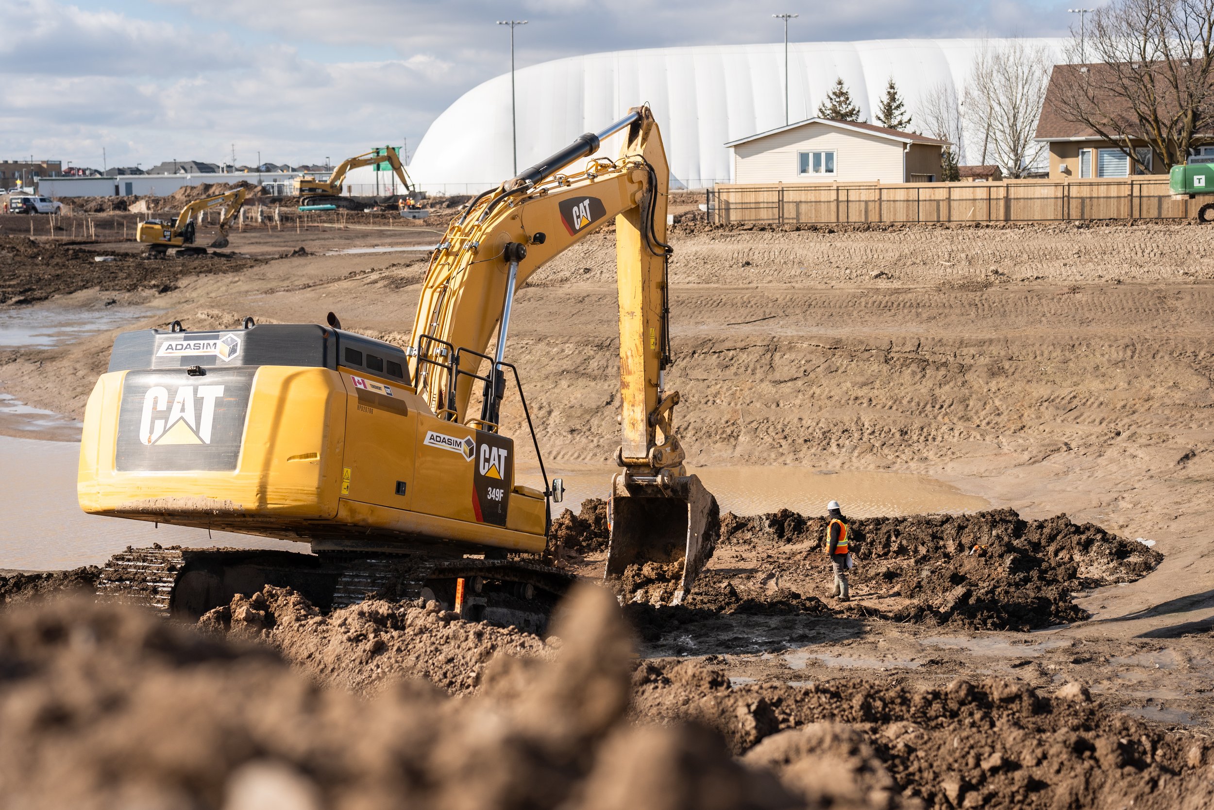 Construction site with excavator digging into muddy ground, worker wearing hard hat and safety vest nearby, and a large white dome structure in the background.