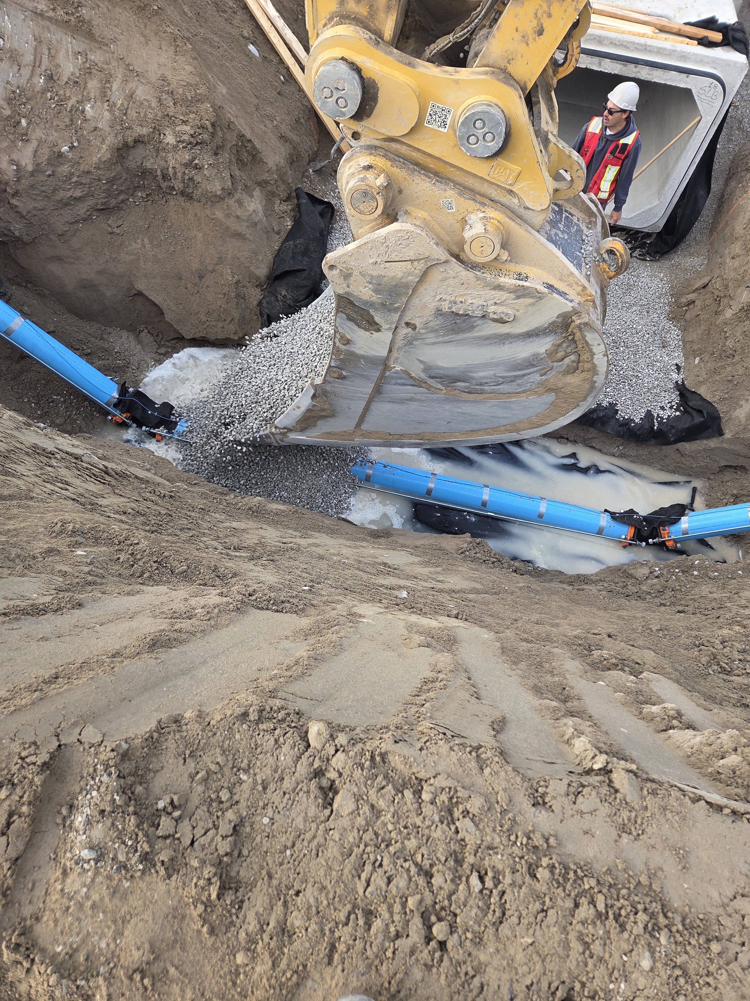 Construction worker operating an excavator digging a trench, with blue pipelines installed in the trench and soil on the surface.