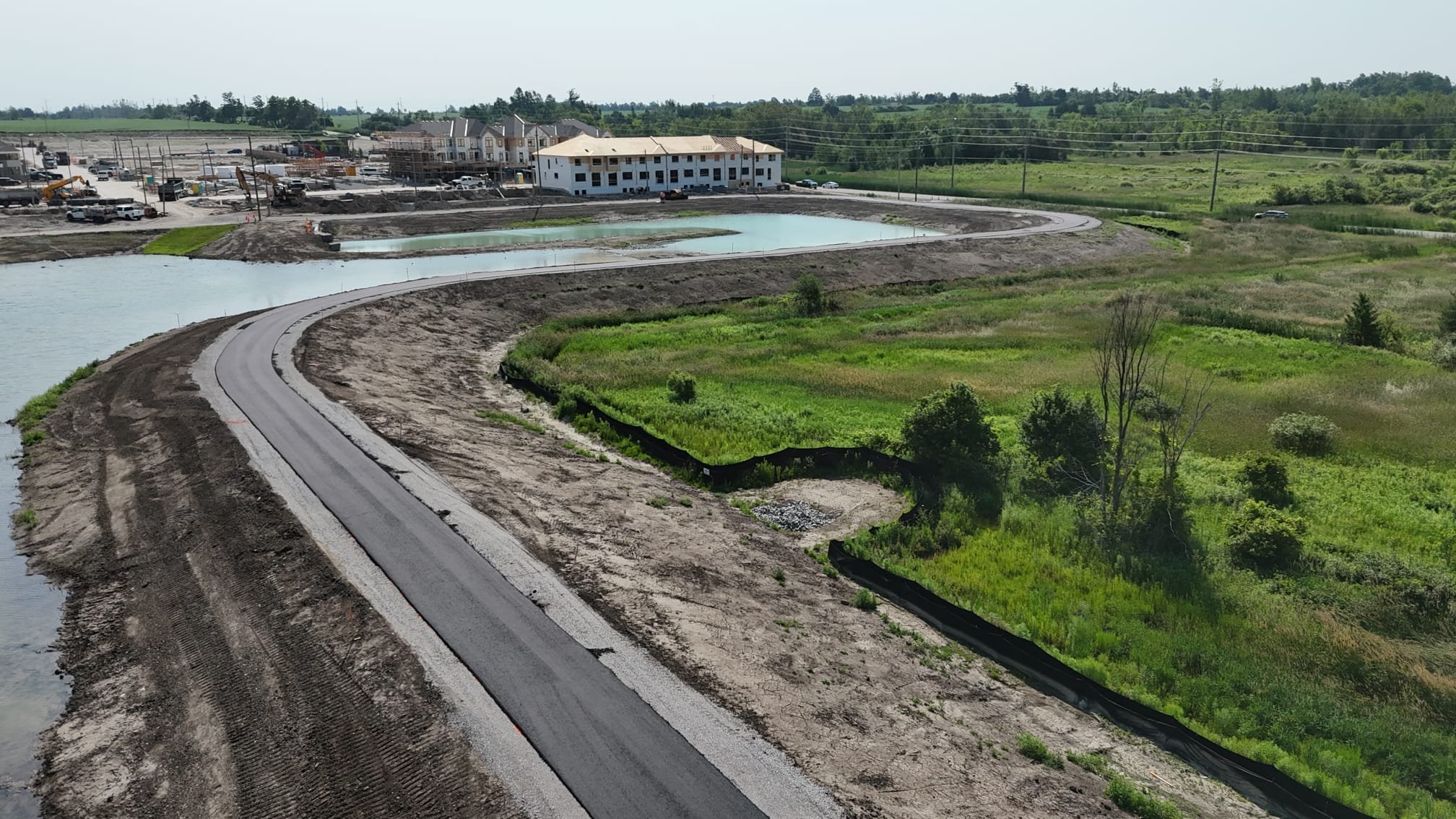 Construction site with a curved road, a pond, and some greenery in the foreground. Buildings and construction activity in the background.