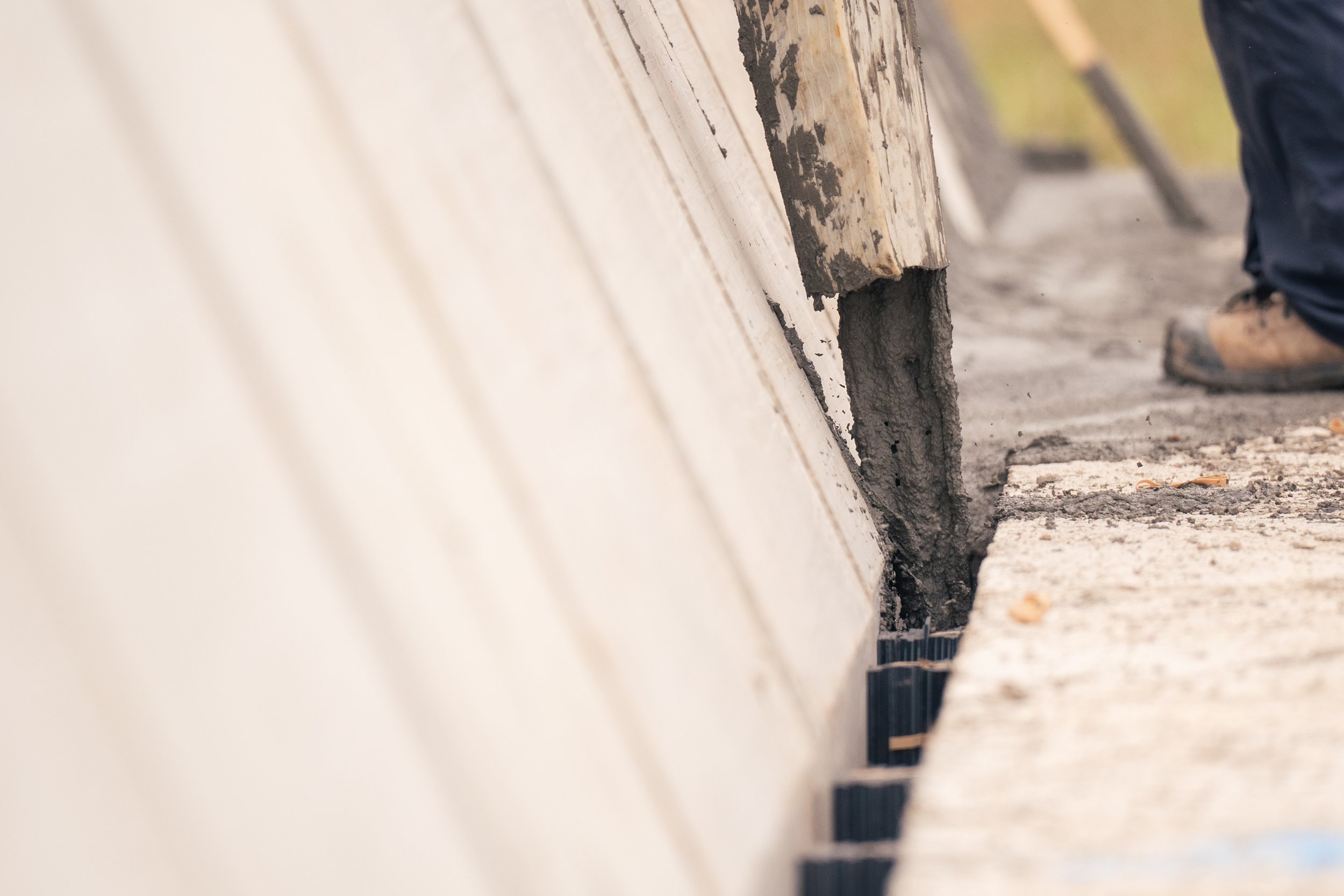 A construction worker laying concrete along the base of a building, with a close-up of the concrete being poured into a slit in the ground.