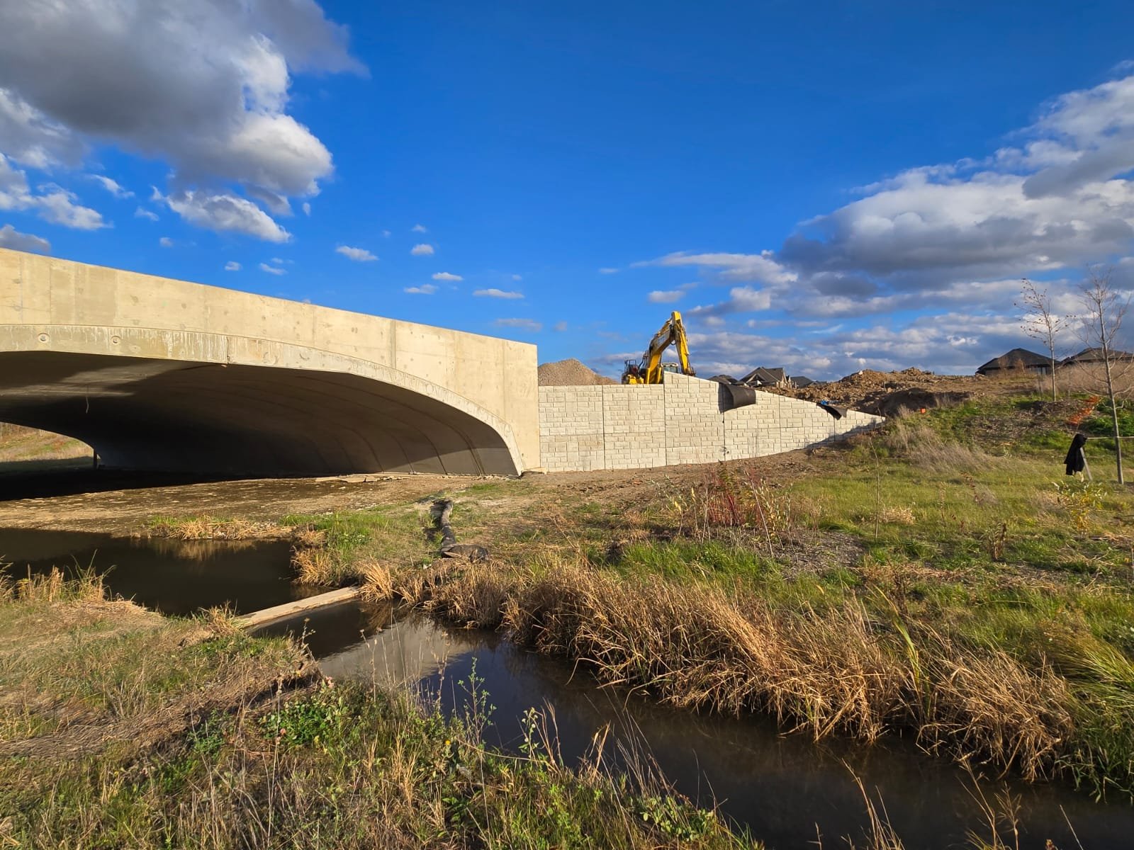 Construction site where a concrete bridge is being built, with a yellow excavator on top of a retaining wall, under a partly cloudy blue sky.
