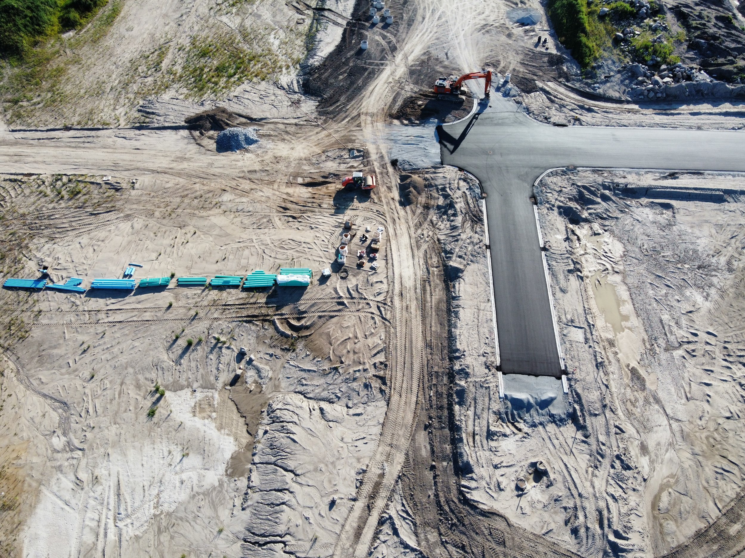 An aerial view of a road construction site showing a new paved road intersection, construction vehicles, and building materials on a cleared, sandy terrain.
