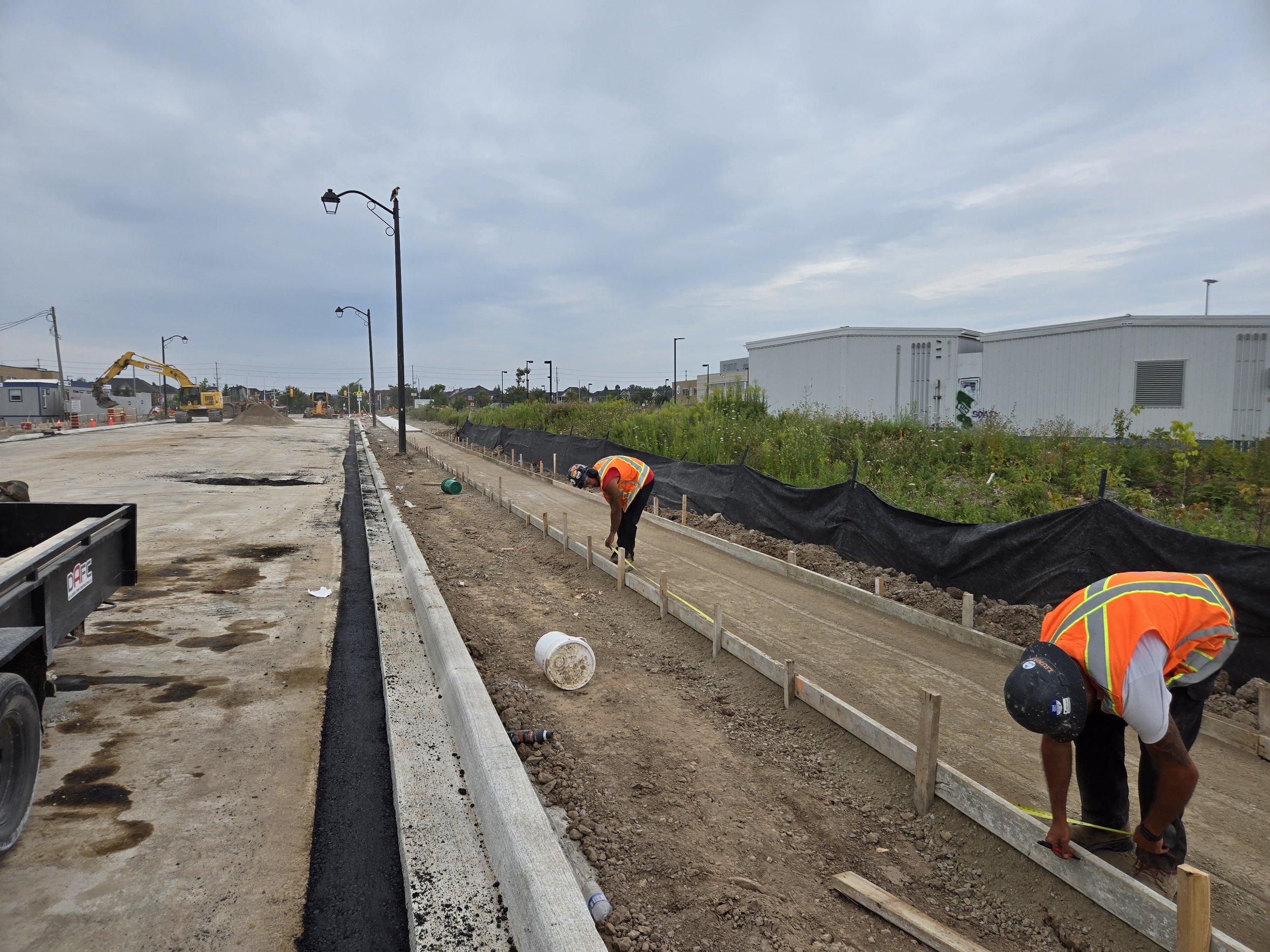 Construction workers measuring and preparing a sidewalk edge on a road construction site, with machinery and partially completed road in the background.