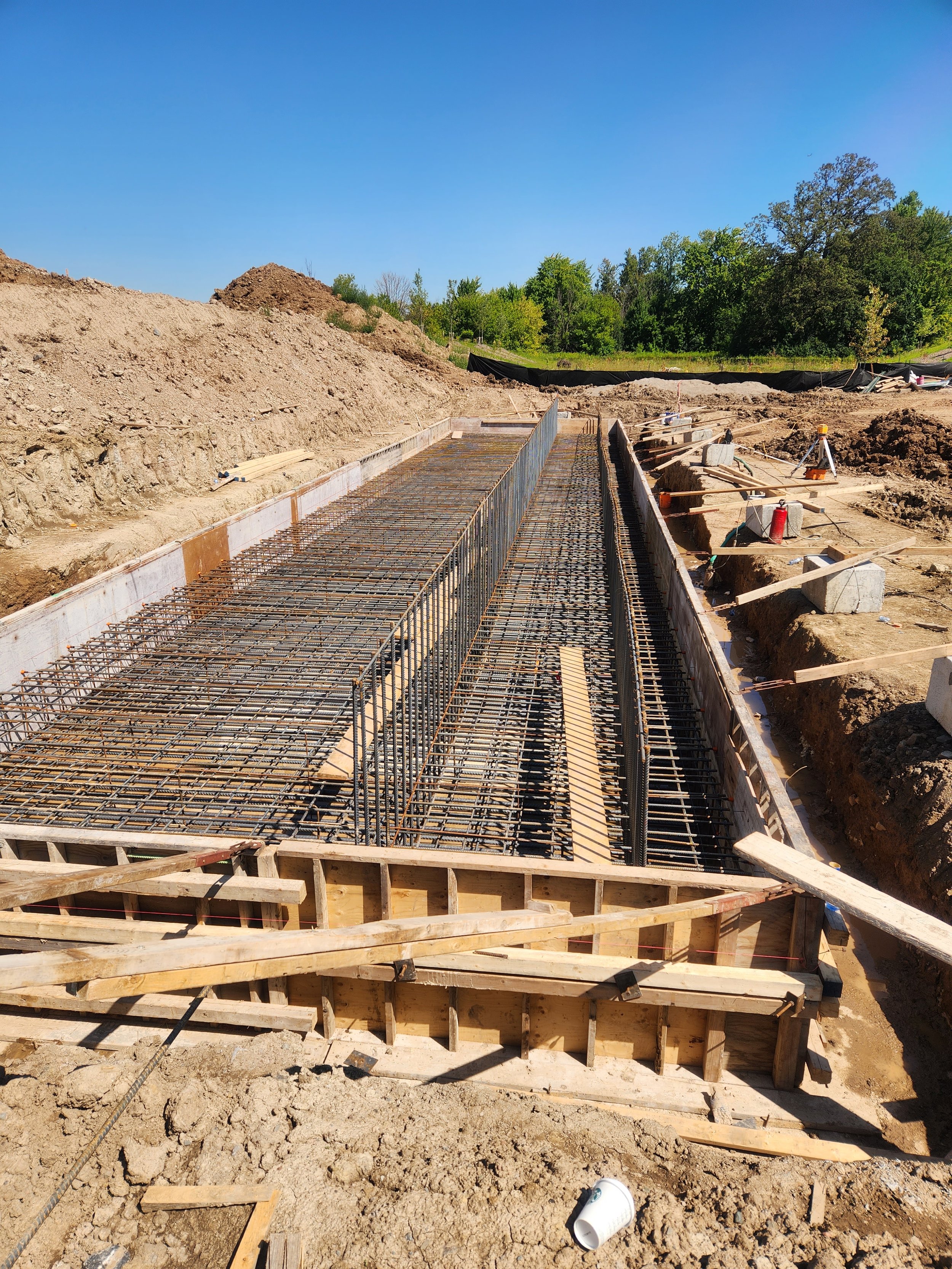 Construction site with reinforced concrete foundation and wooden framework, surrounded by dirt and construction tools under a clear blue sky.