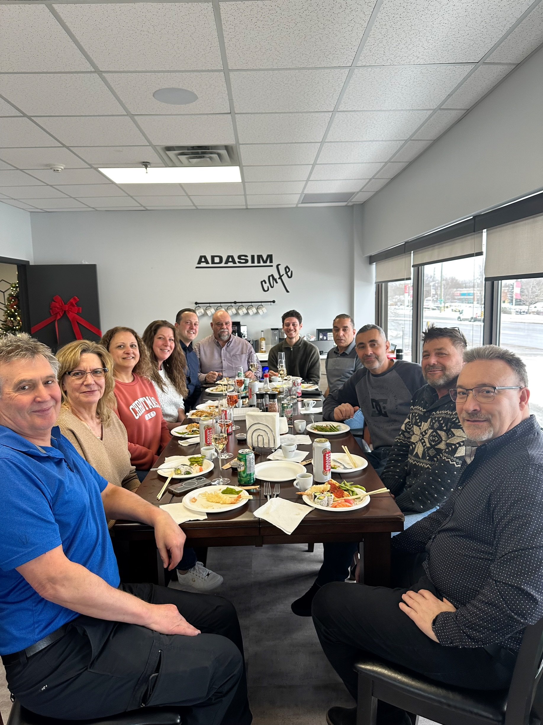 A group of fourteen people sitting at a long dining table in a cafe, having a meal together. The cafe has large windows with blinds, and a sign on the wall reads 'ADASIM cafe.' Plates with food, drinks, and condiments are on the table.
