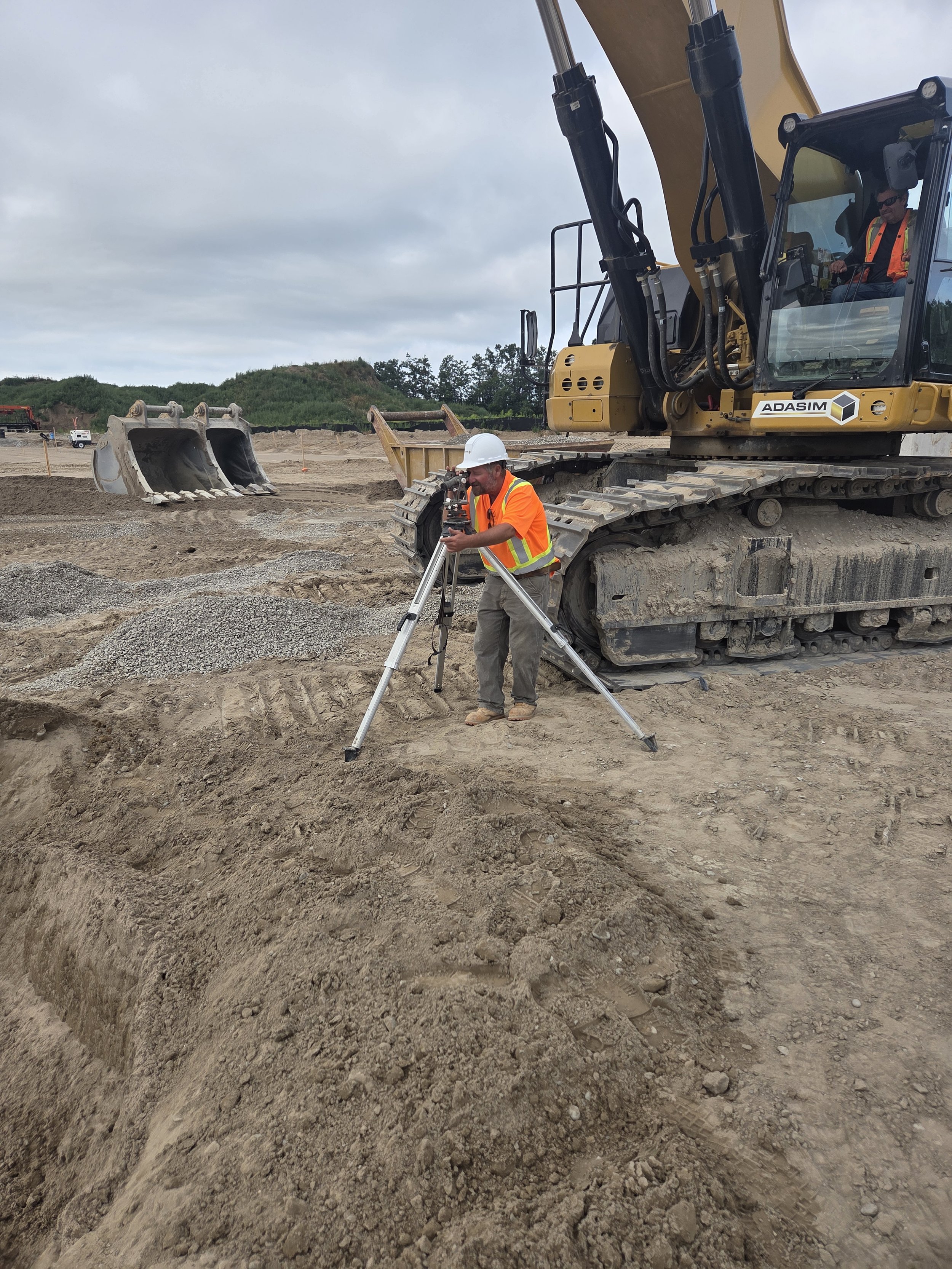 Construction site with a person in an orange safety vest and white hard hat operating a theodolite, and another person on an excavator in the background.