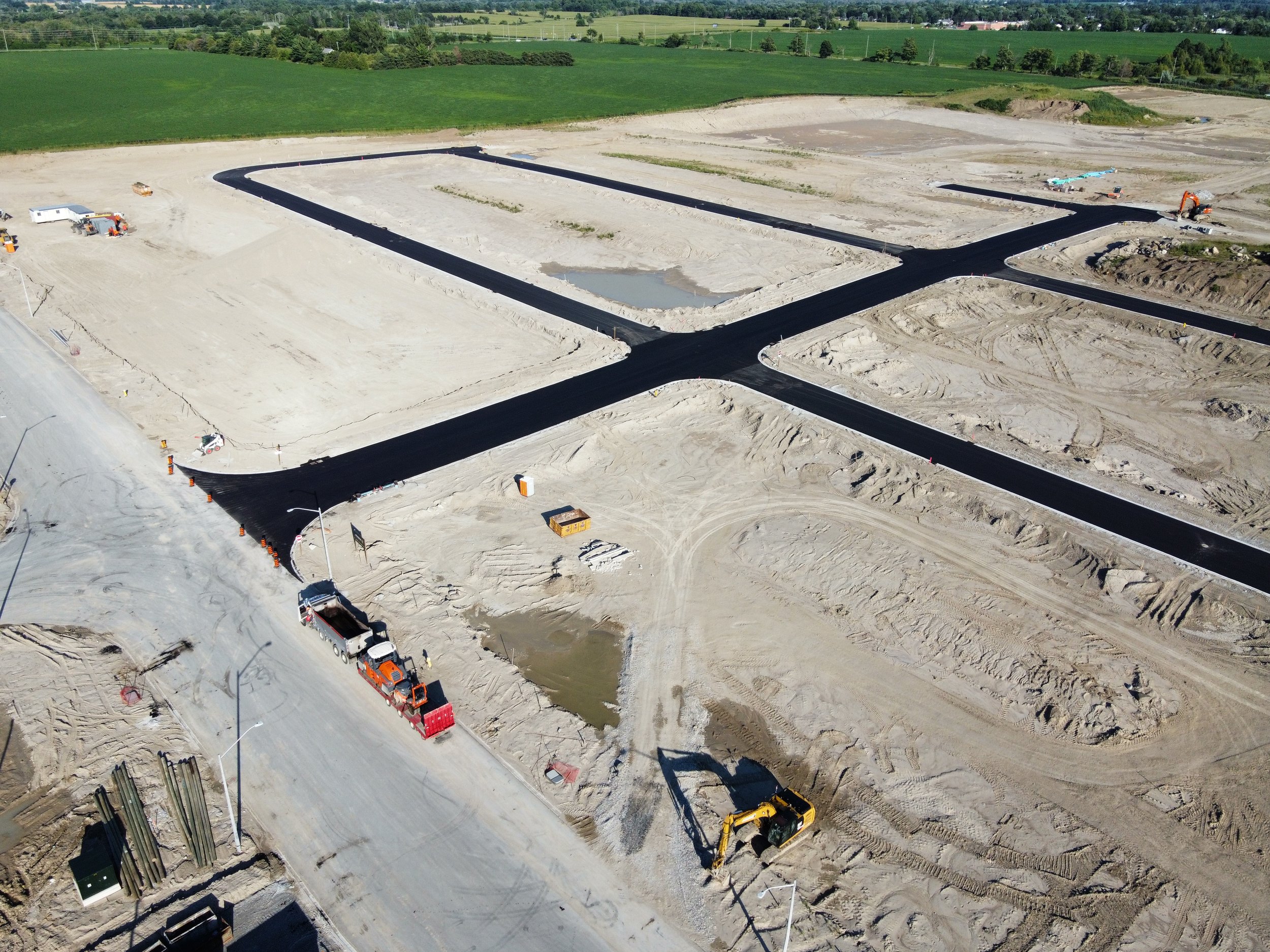 Aerial view of a construction site with freshly paved roads forming a grid pattern, with construction vehicles and equipment, excavated areas, and surrounding fields.